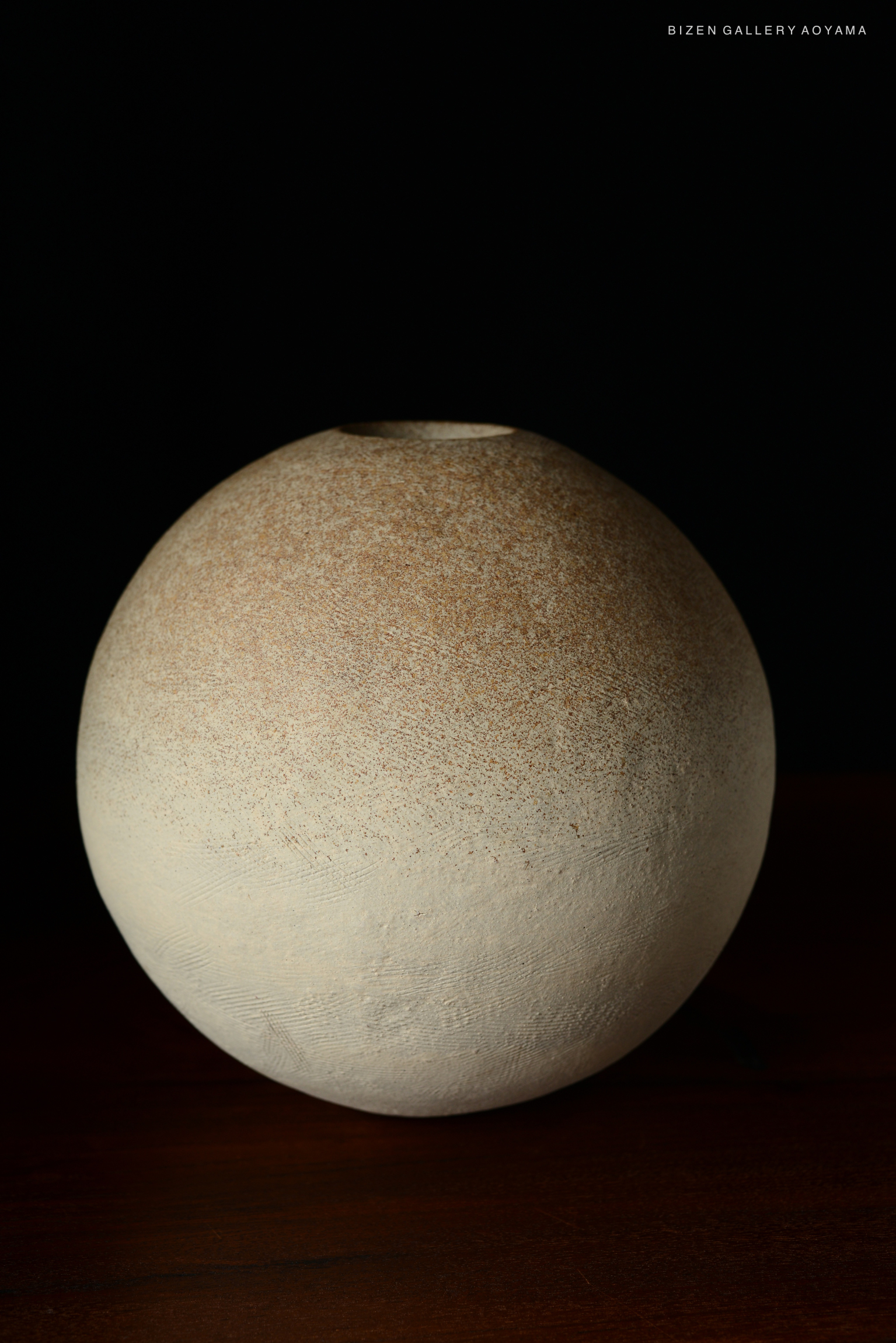 A spherical Bizen pottery vase with a smooth, light-colored surface and subtle textures, presented against a dark background.