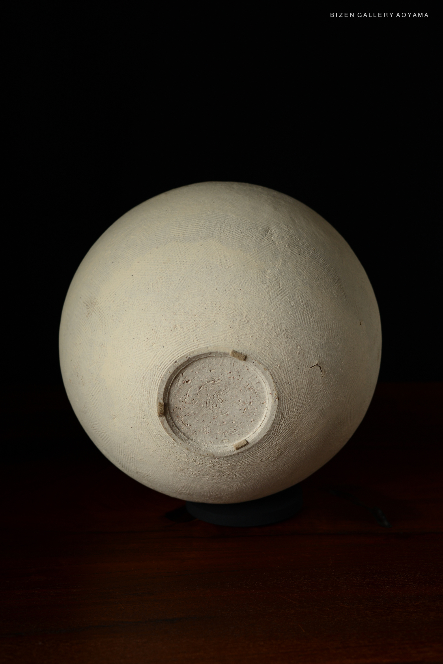A close-up view of the base of a round Bizen pottery vase, showcasing its textured surface and markings. The vase is positioned against a dark background.