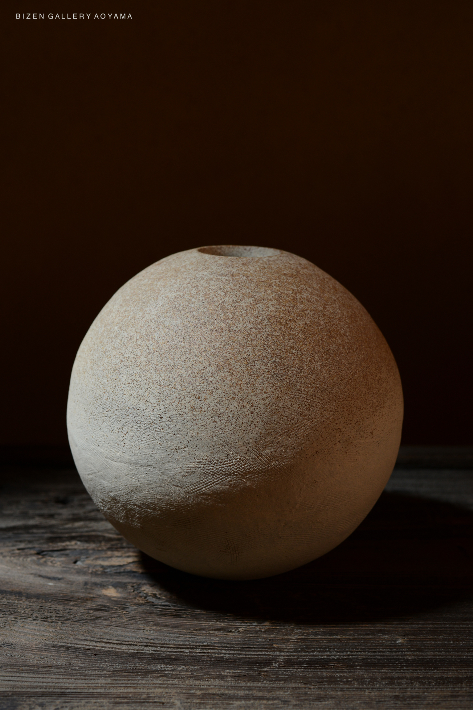 A round, textured Bizen pottery vase with a neutral color, set against a dark background.