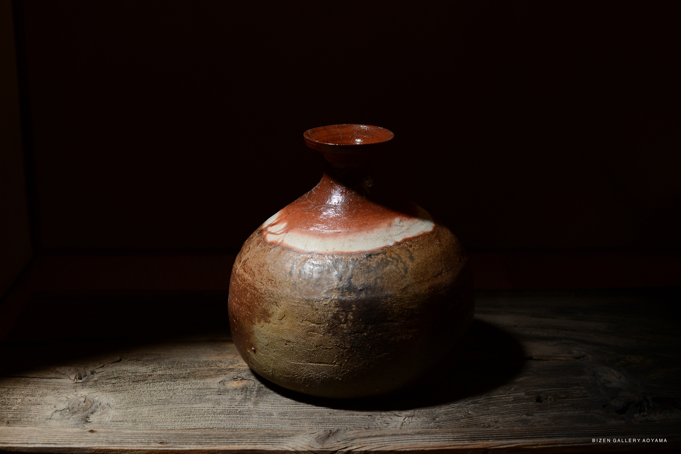 A traditional Bizen pottery Tokkuri with a rounded body and a narrow neck, displayed on a wooden surface in a dimly lit environment.