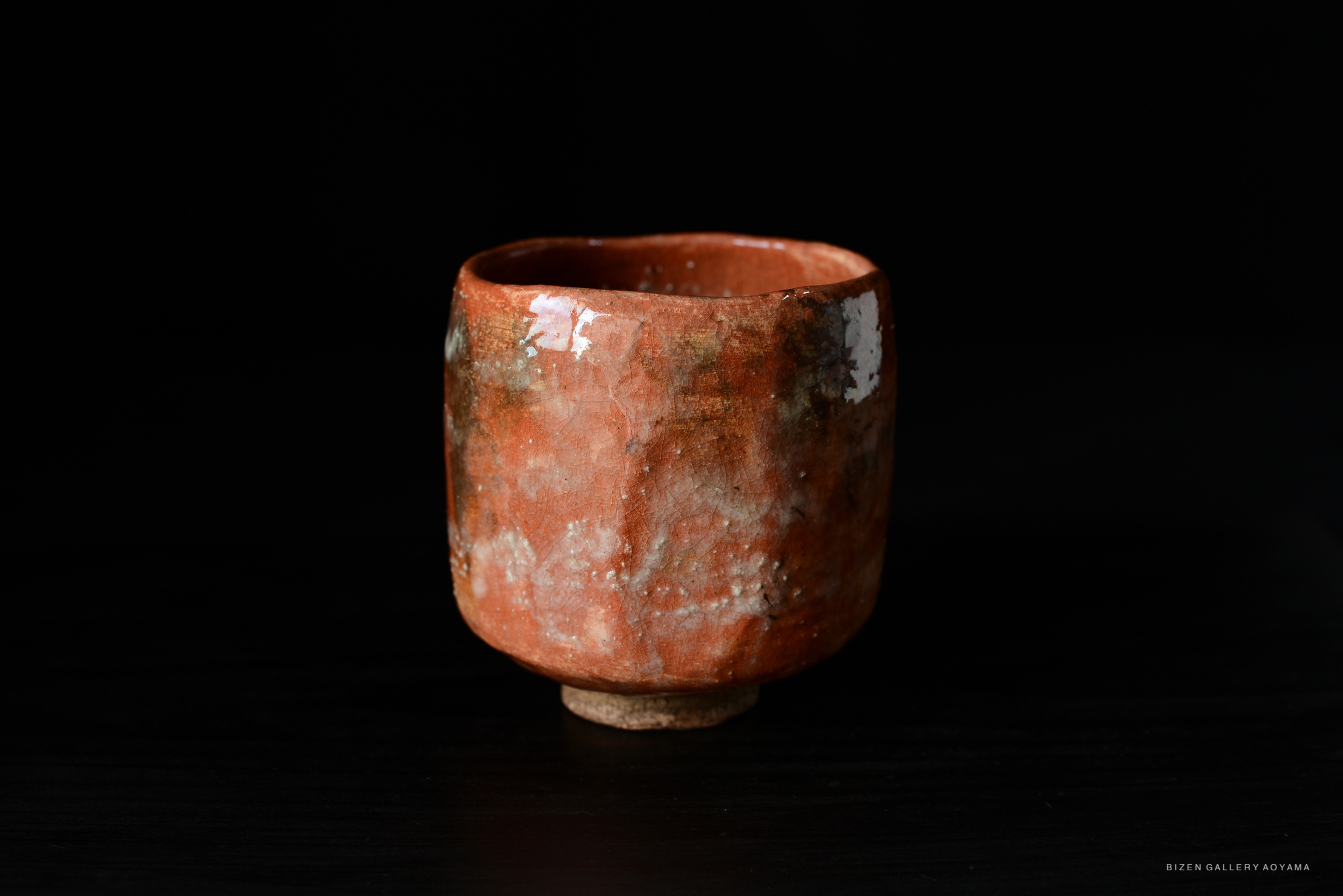 A handcrafted red ceramic tea bowl with a textured surface, displayed against a black background, highlighting its unique shape and artistic details.