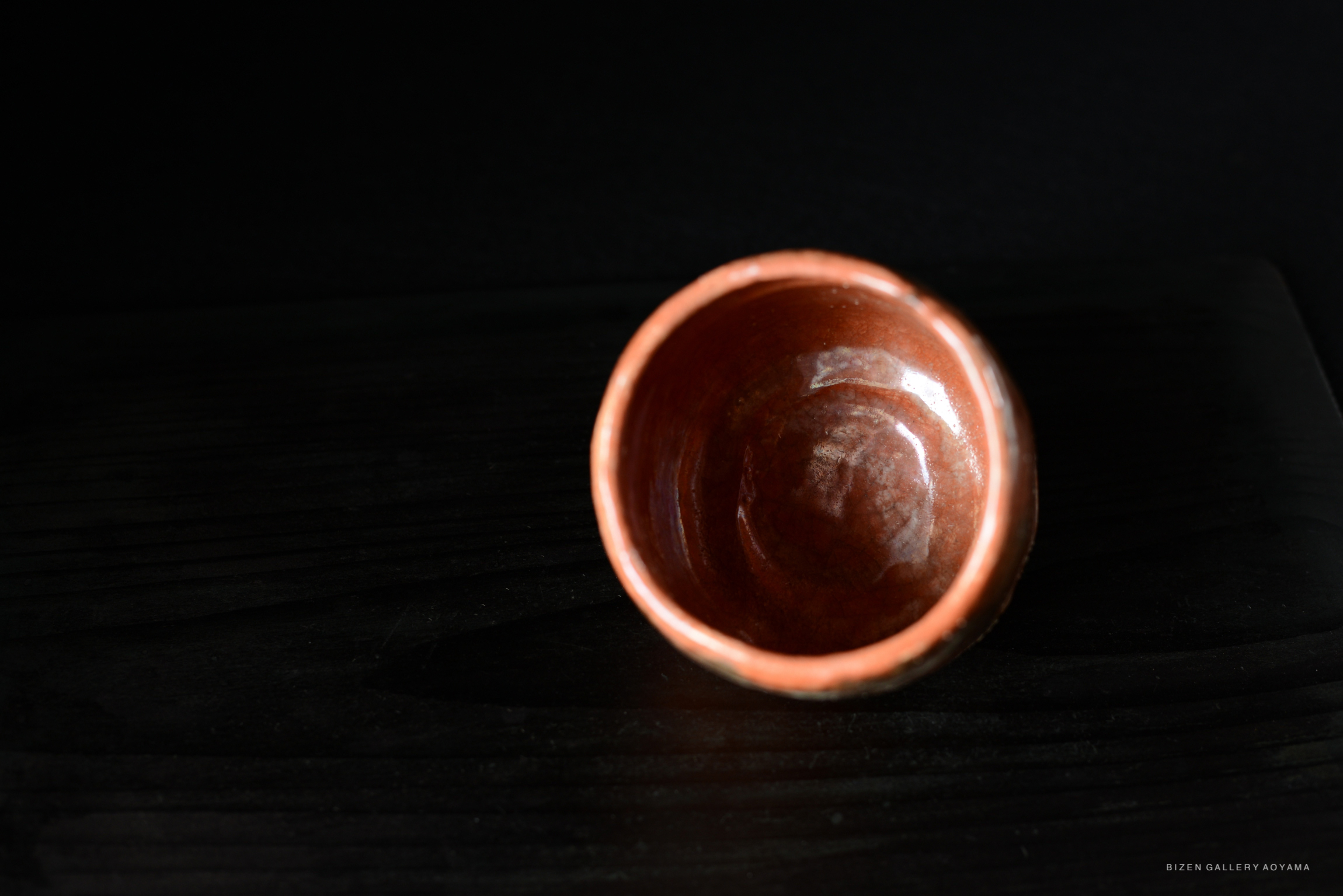 A close-up view of a red ceramic tea bowl with a smooth, shiny interior and a textured exterior, placed on a dark wooden surface.