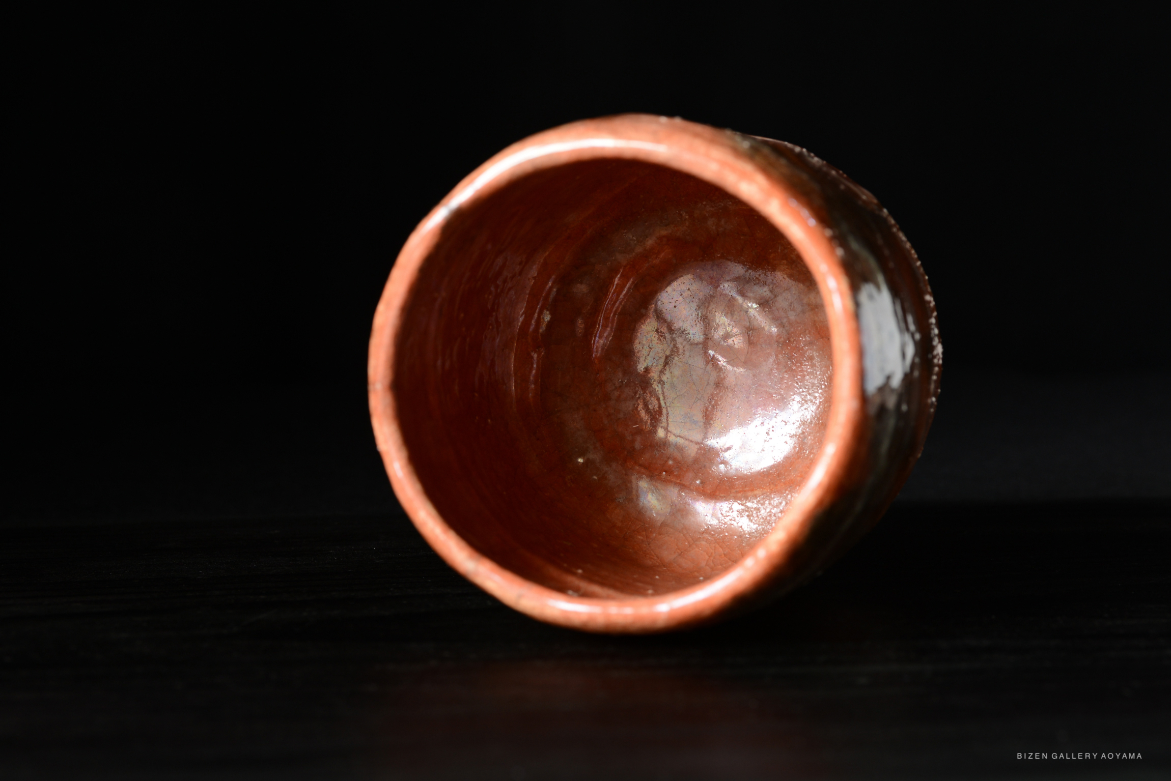 A close-up view of a red tea bowl with a smooth, glossy interior and a slightly rough exterior, set against a dark background.