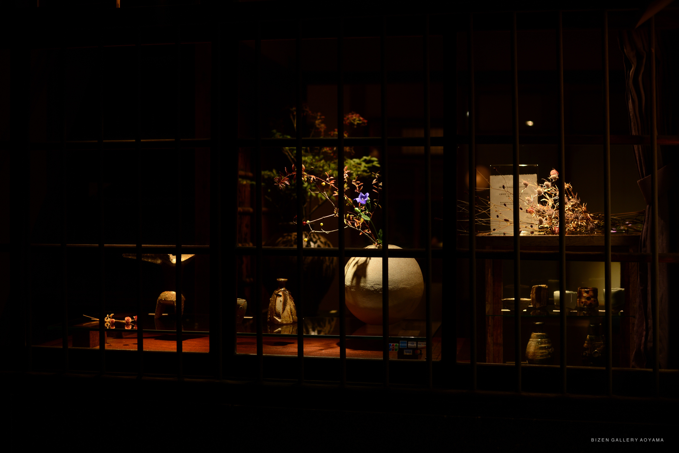 A window display showcasing traditional Bizen pottery and floral arrangements in a dimly lit setting.