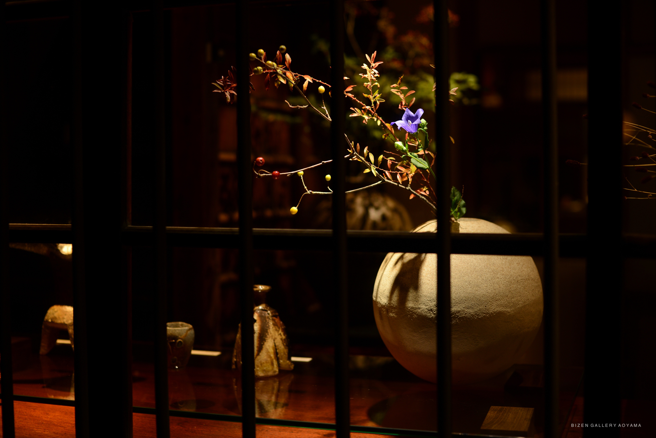 A beautifully arranged flower display inside a wooden showcase, featuring a round Bizen pottery vase with various branches and a purple flower illuminated by soft lighting.