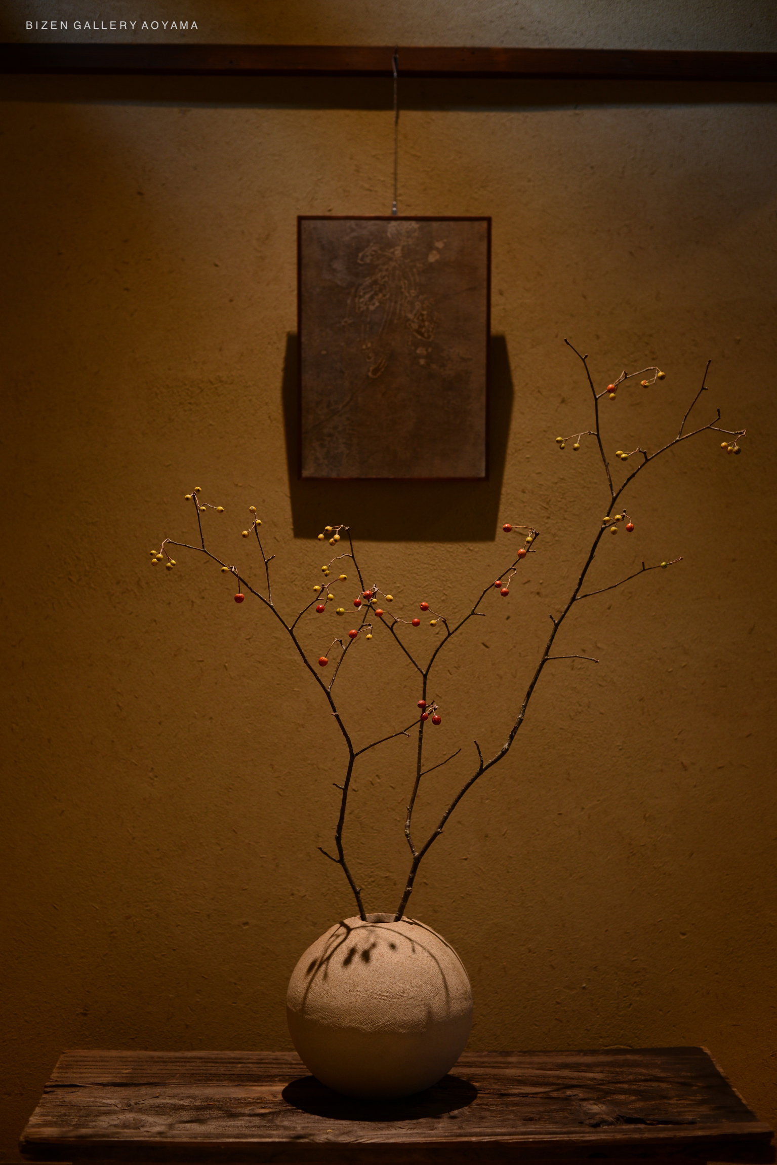 A decorative Bizen vase with twigs featuring small berries, placed on a wooden table against a textured wall, with an artwork hanging above.