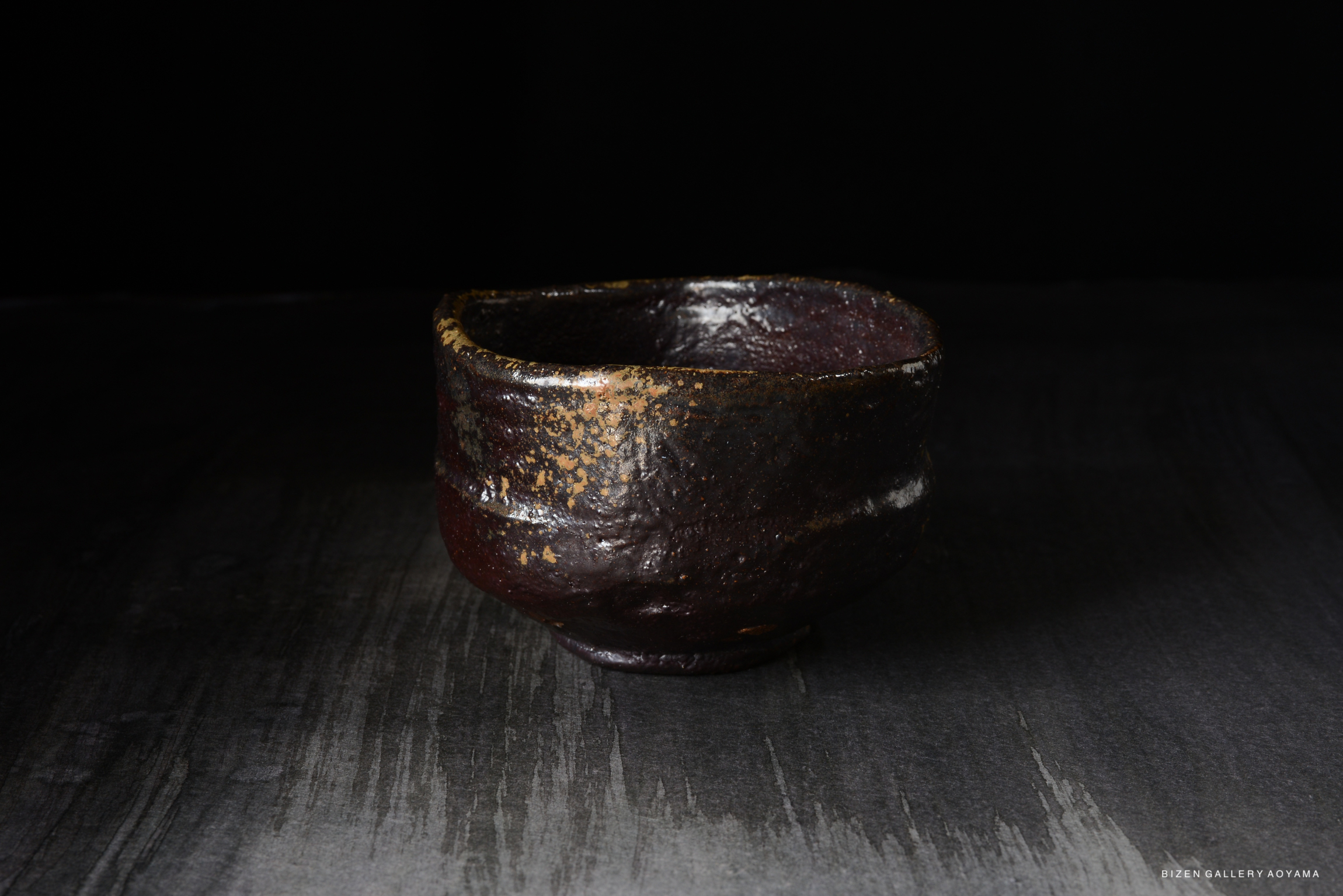 A handcrafted dark ceramic tea bowl with a slightly irregular shape, displaying a textured surface and subtle gold accents, set against a dark background.