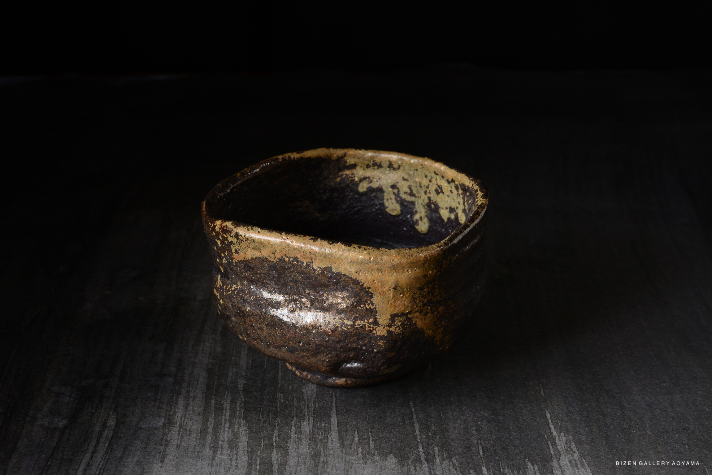 A rustic, handcrafted ceramic bowl with a dark, textured surface and a contrasting lighter rim, positioned on a dark background.