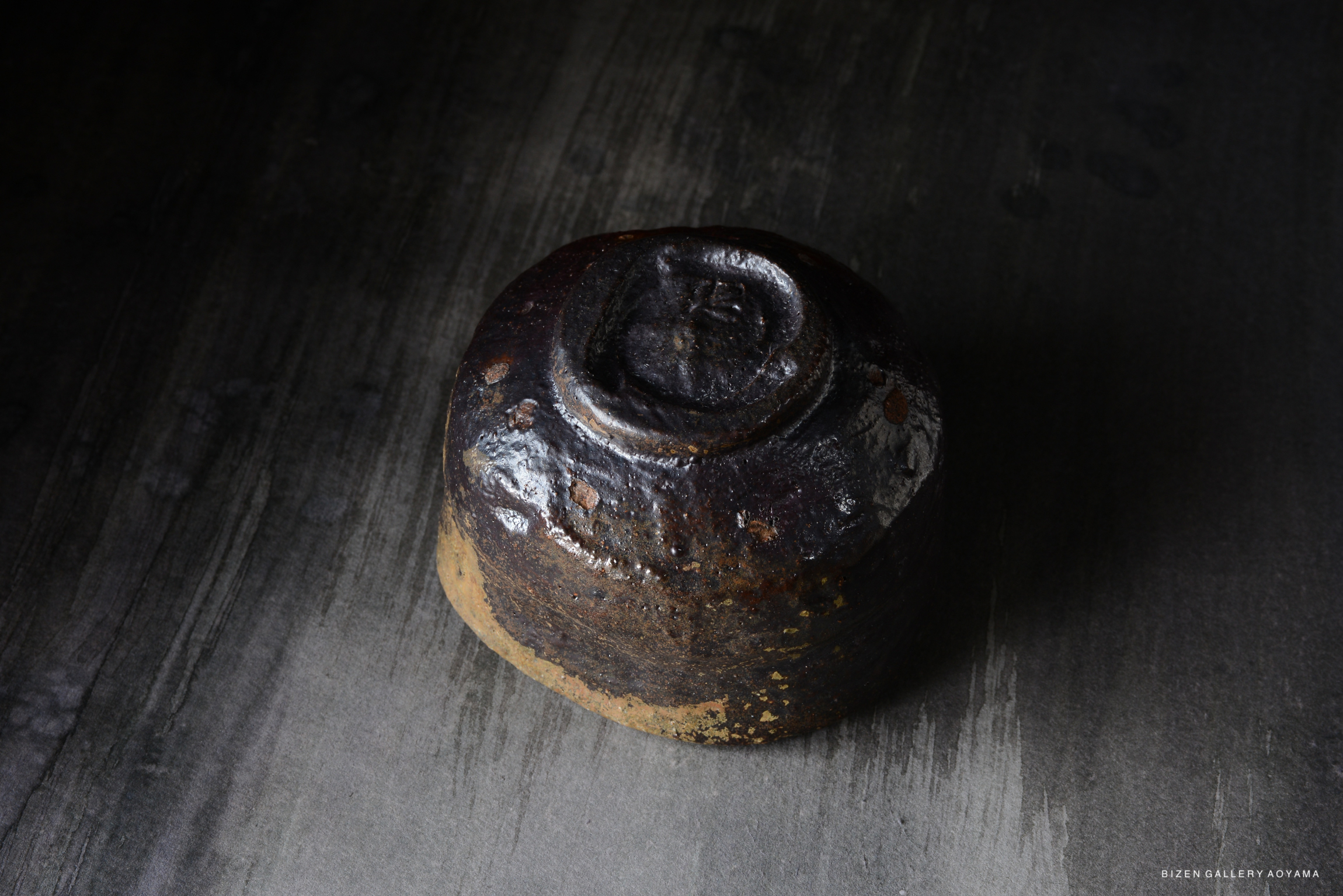 A close-up image of a traditional Bizen pottery piece with a textured, dark surface, sitting on a dark wooden table.