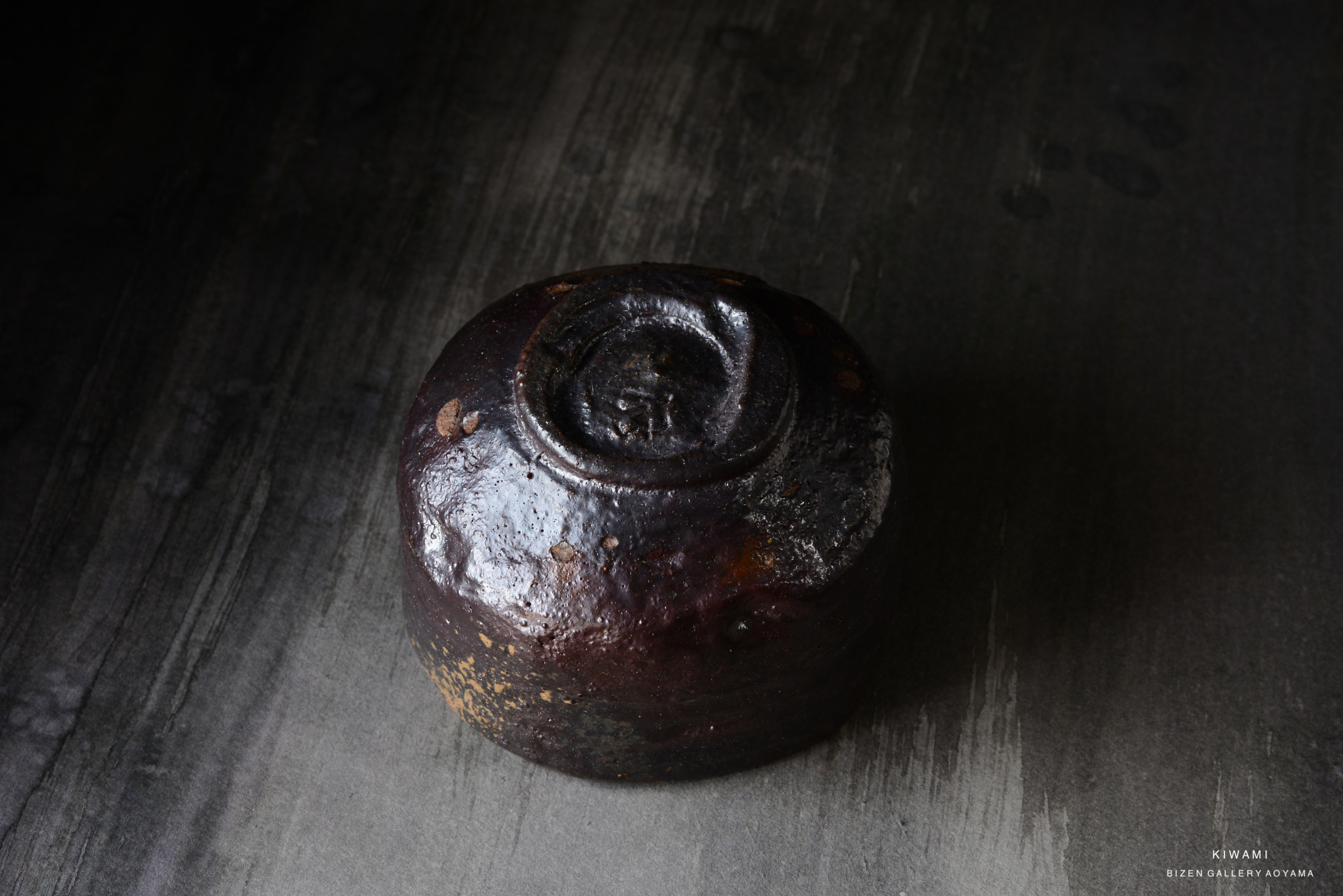 A top view of a rustic, dark ceramic teapot with a rounded body and a textured surface, placed on a dark wooden table.