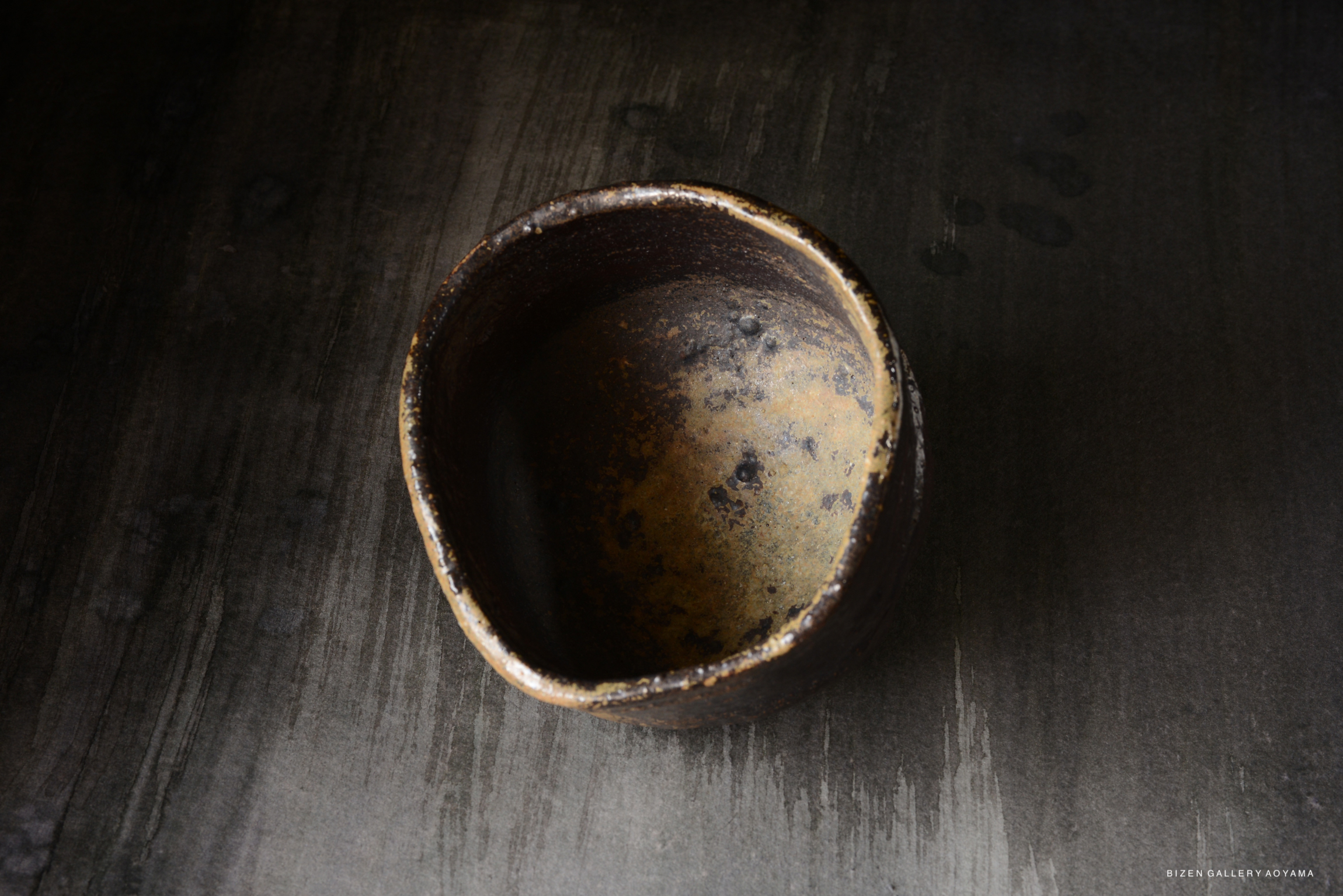 A top-down view of a rustic, dark ceramic bowl with a textured surface and slight wear, set against a dark background.