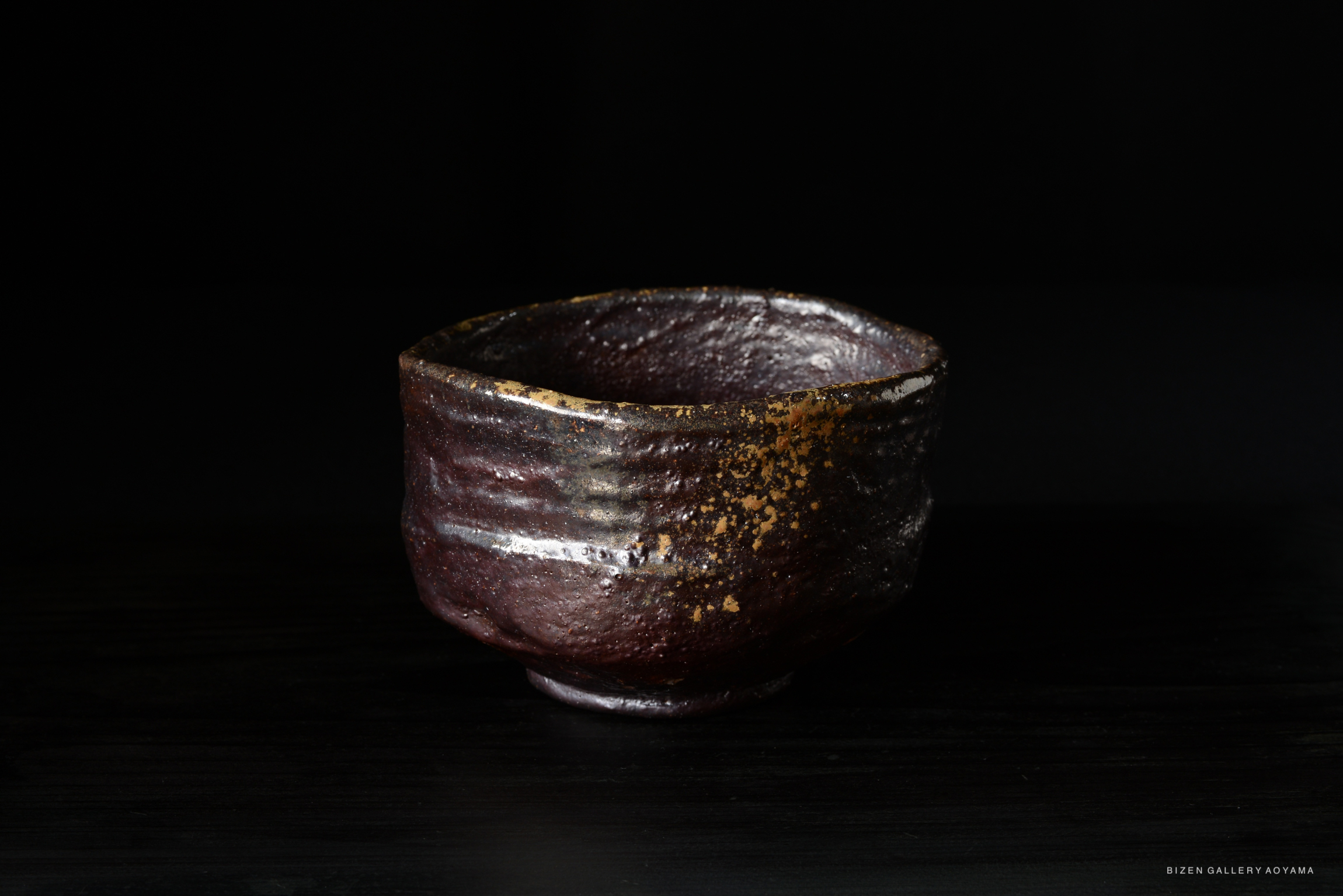 A close-up view of a textured, dark ceramic tea bowl with a rustic finish, displayed against a black background.