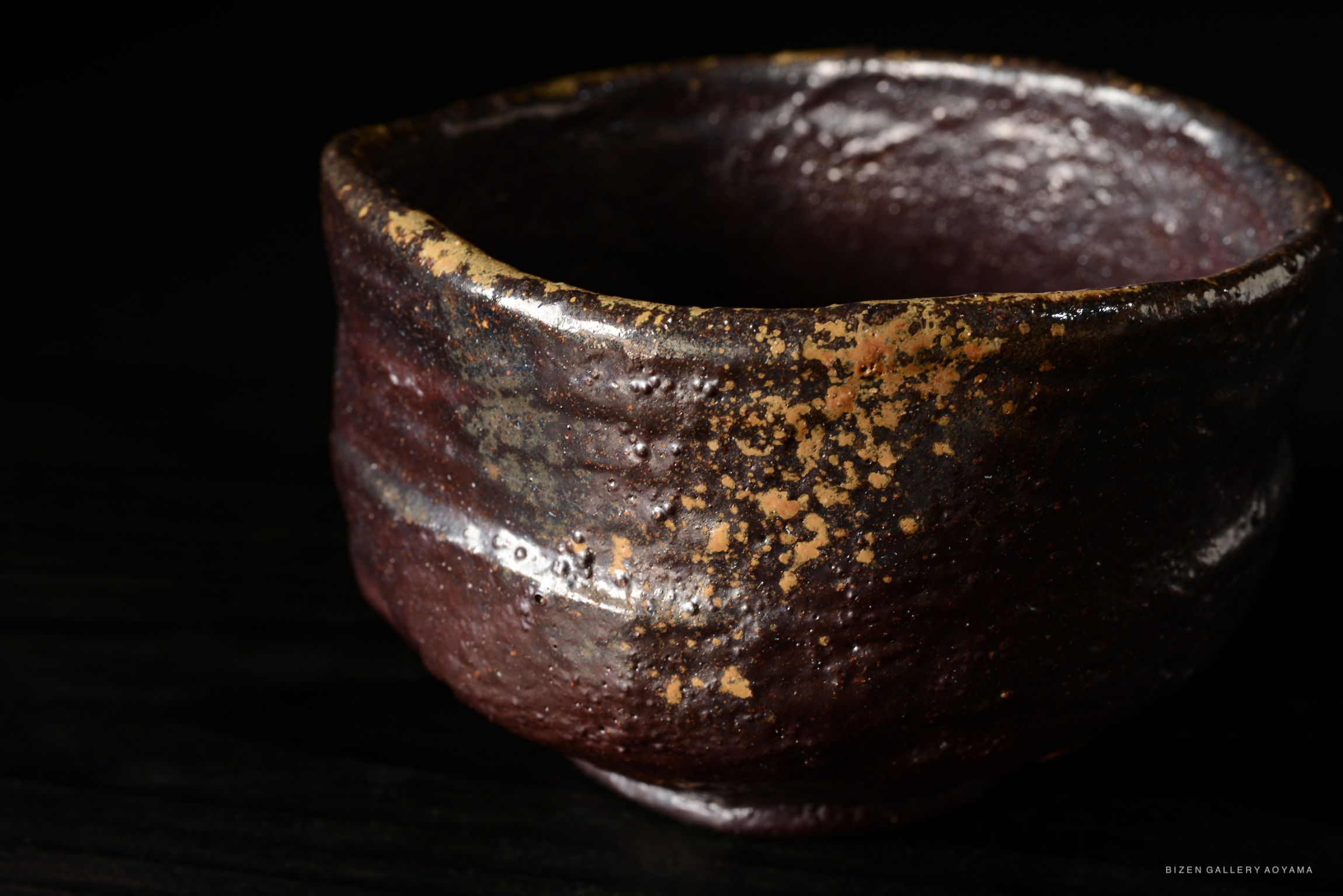 Close-up of a textured ceramic bowl with a dark, earthy glaze and speckles of golden color against a black background.
