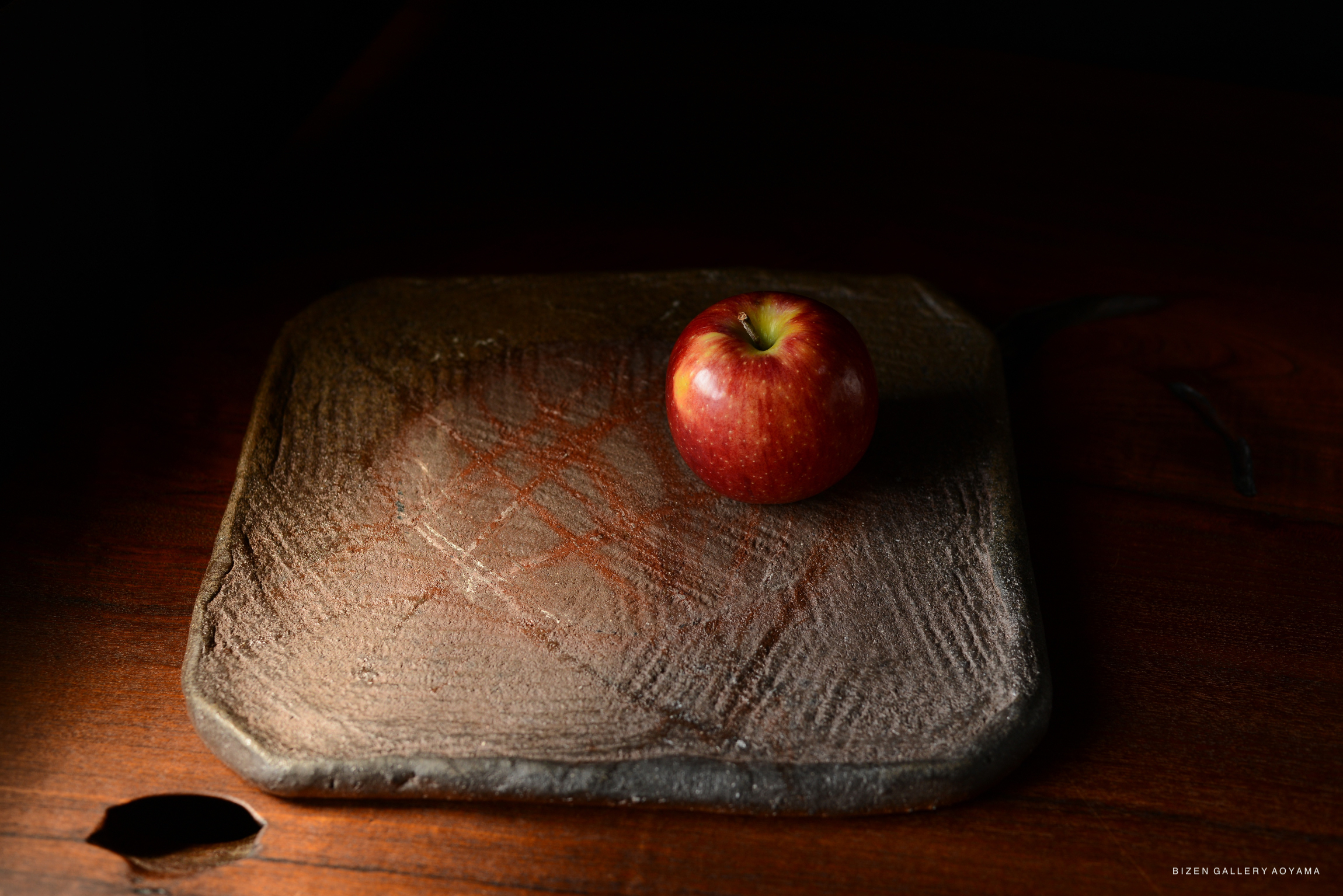 A red apple placed on a textured, rustic square plate resting on a wooden surface.