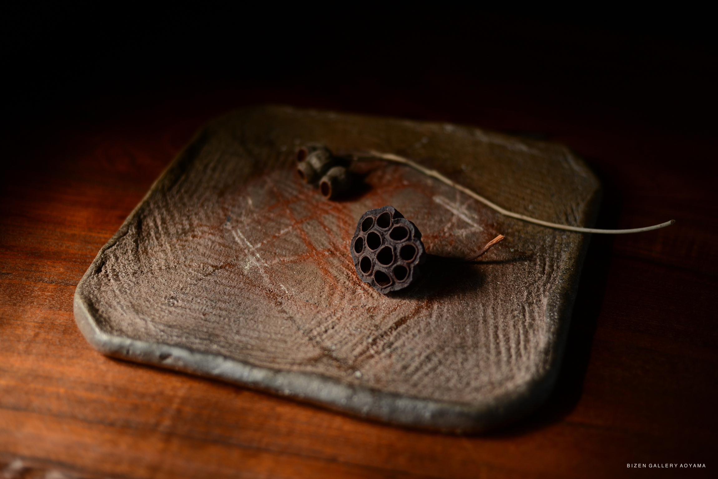 A decorative arrangement featuring a dried lotus seed pod and additional natural elements atop a textured, clay plate on a wooden surface.
