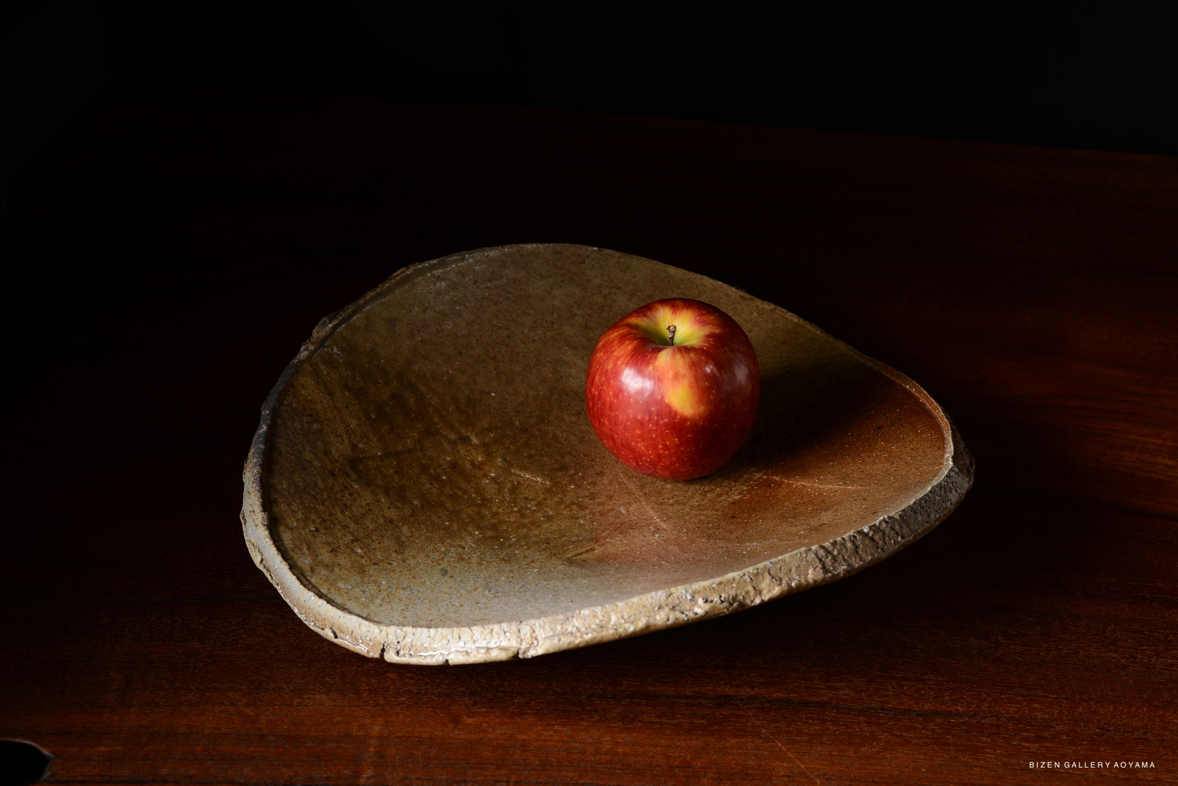 A vibrant red apple sitting on a unique, rustic ceramic plate against a dark background.