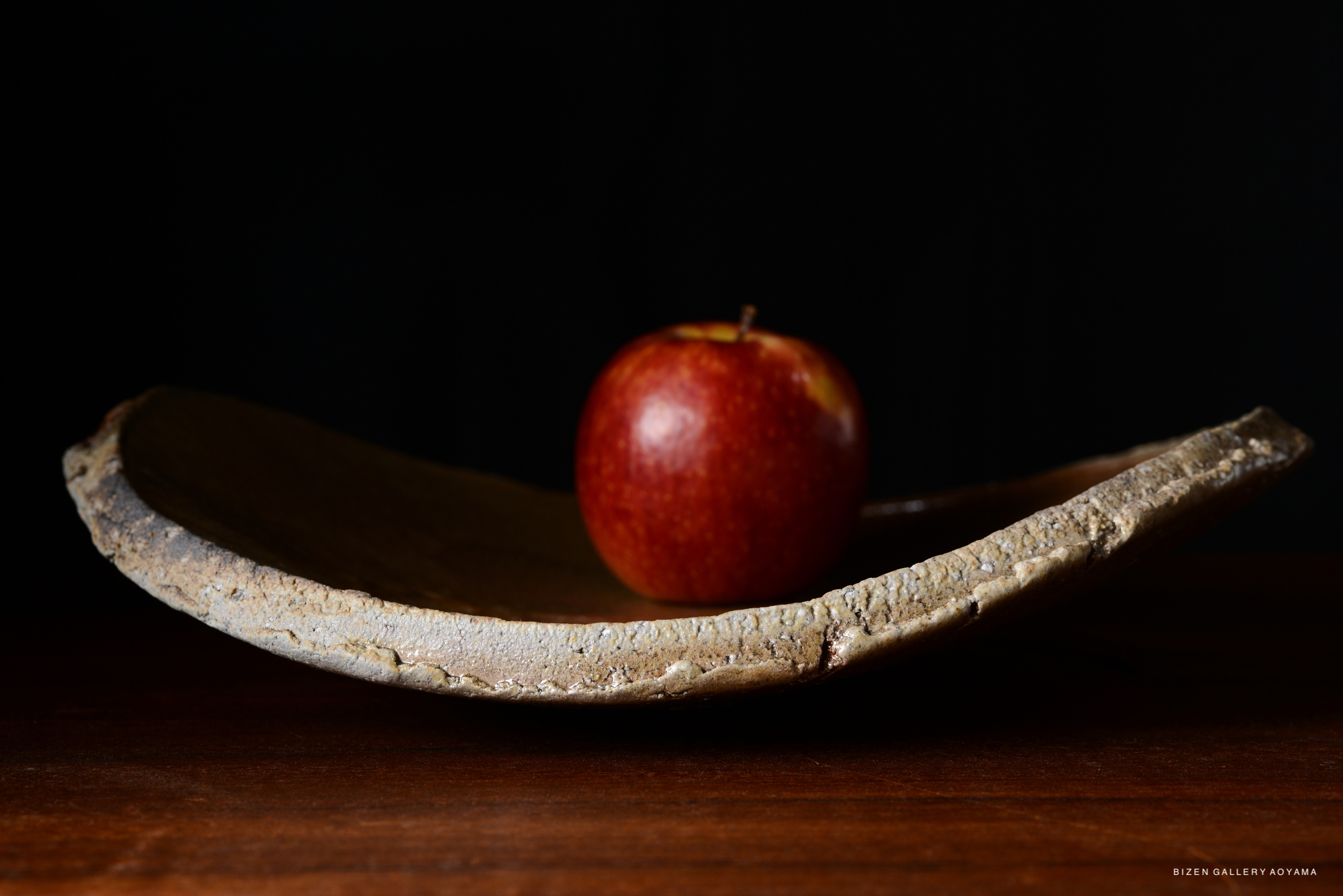 A close-up image of a polished wooden dish with a red apple resting on it, set against a dark background.