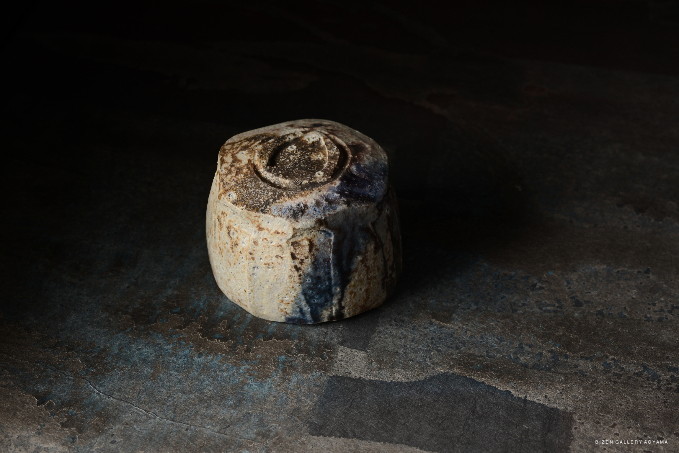 A close-up of a rustic ceramic piece with textured surface and earthy tones, set against a dark background.