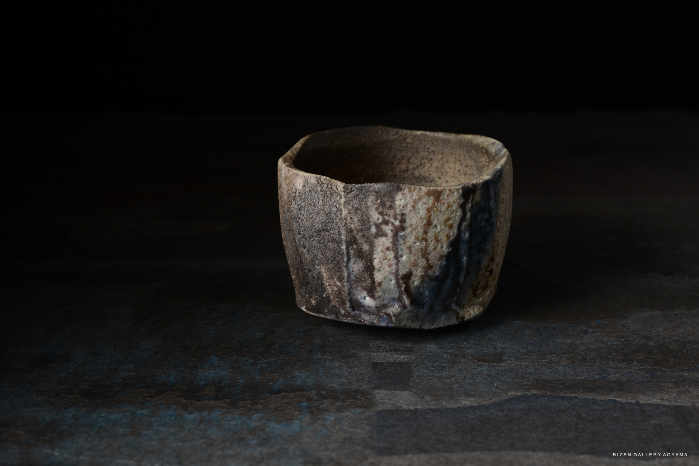 A handcrafted ceramic bowl with an uneven shape and a rustic finish, featuring earthy tones and textures against a dark background.