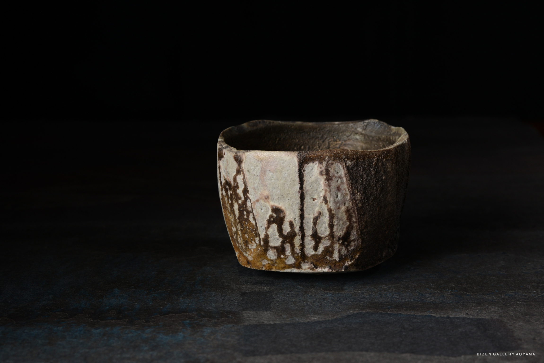A rustic ceramic tea bowl with an uneven, textured surface featuring earthy tones and subtle patterns, displayed on a dark background.