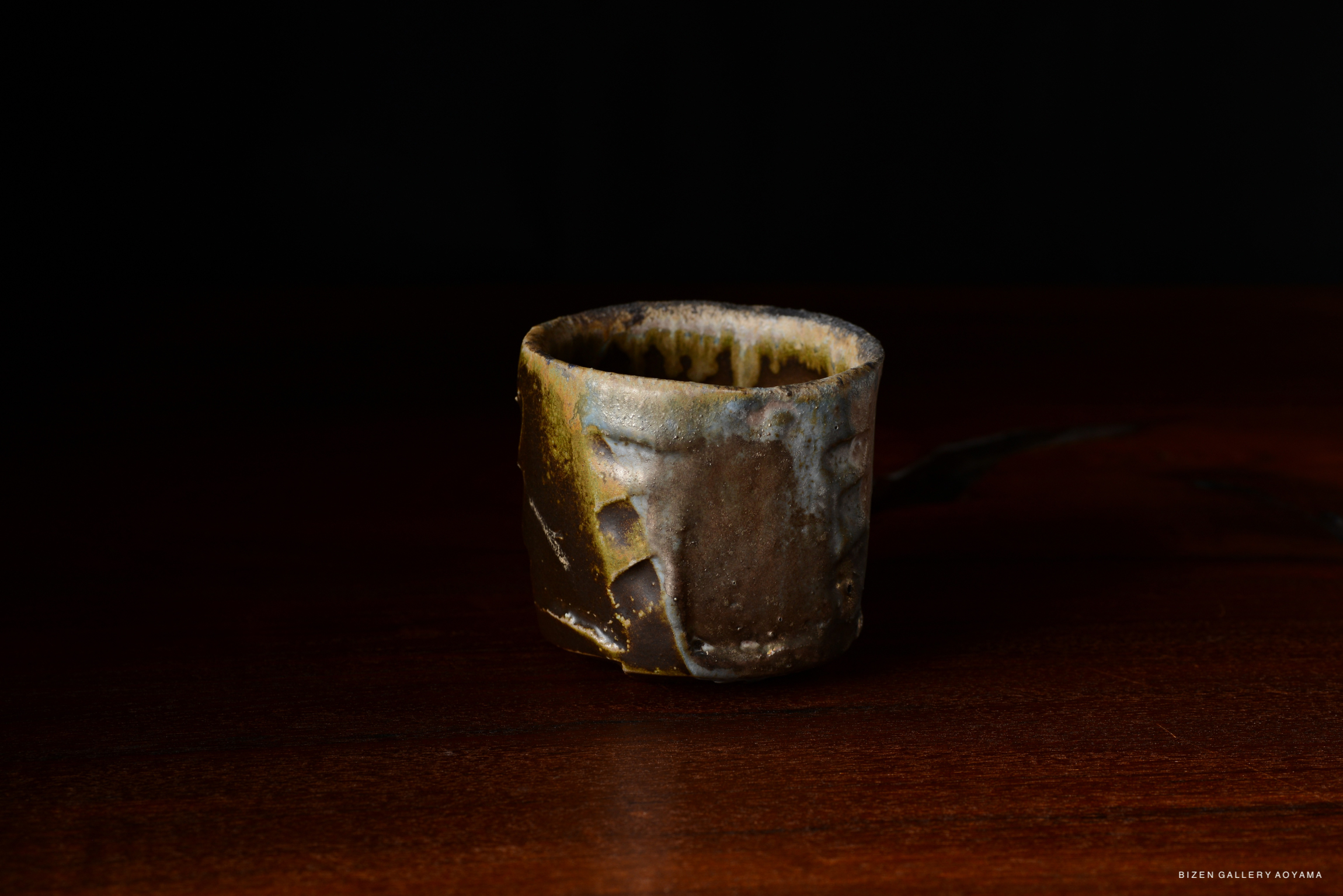A handcrafted ceramic cup with a unique texture and earthy colors, placed on a wooden surface against a dark background.