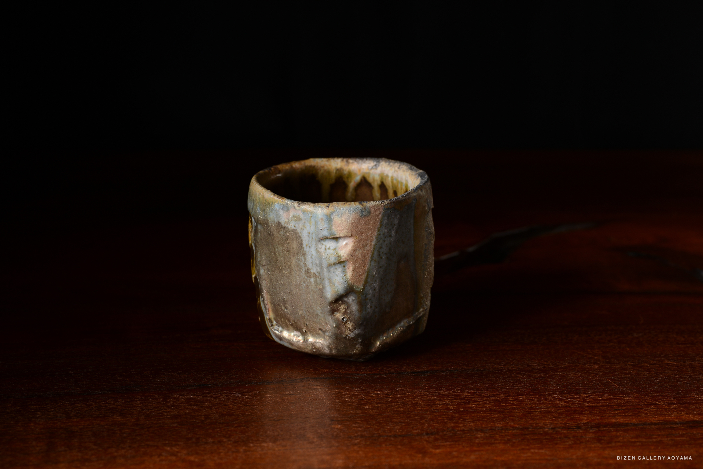A handmade ceramic cup with a unique texture and earthy colors, placed on a wooden surface against a dark background.