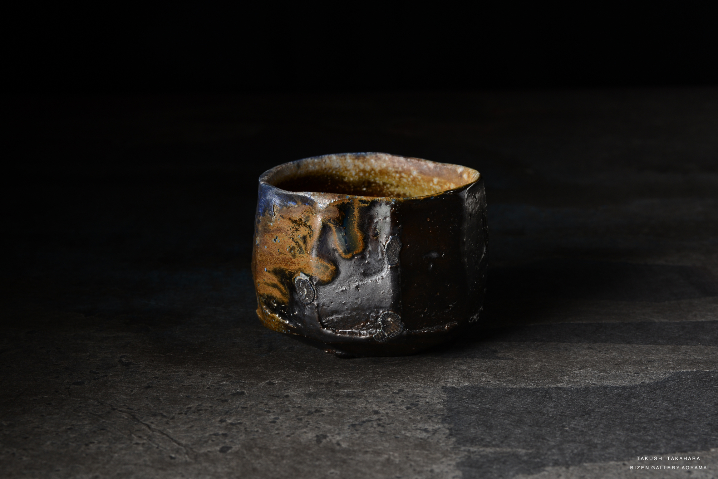 A close-up image of a handcrafted pottery cup, featuring dark black and brown glazes with a textured surface, placed on a dark stone background.