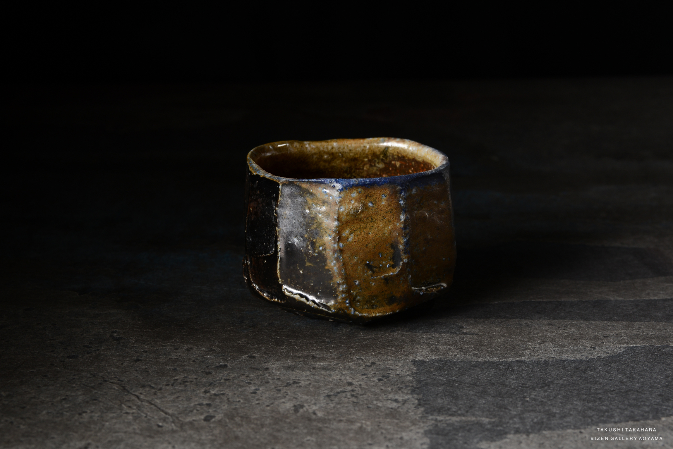 A handcrafted ceramic cup with a unique, textured surface showcasing earthy tones of brown, black, and hints of blue, set on a dark background.