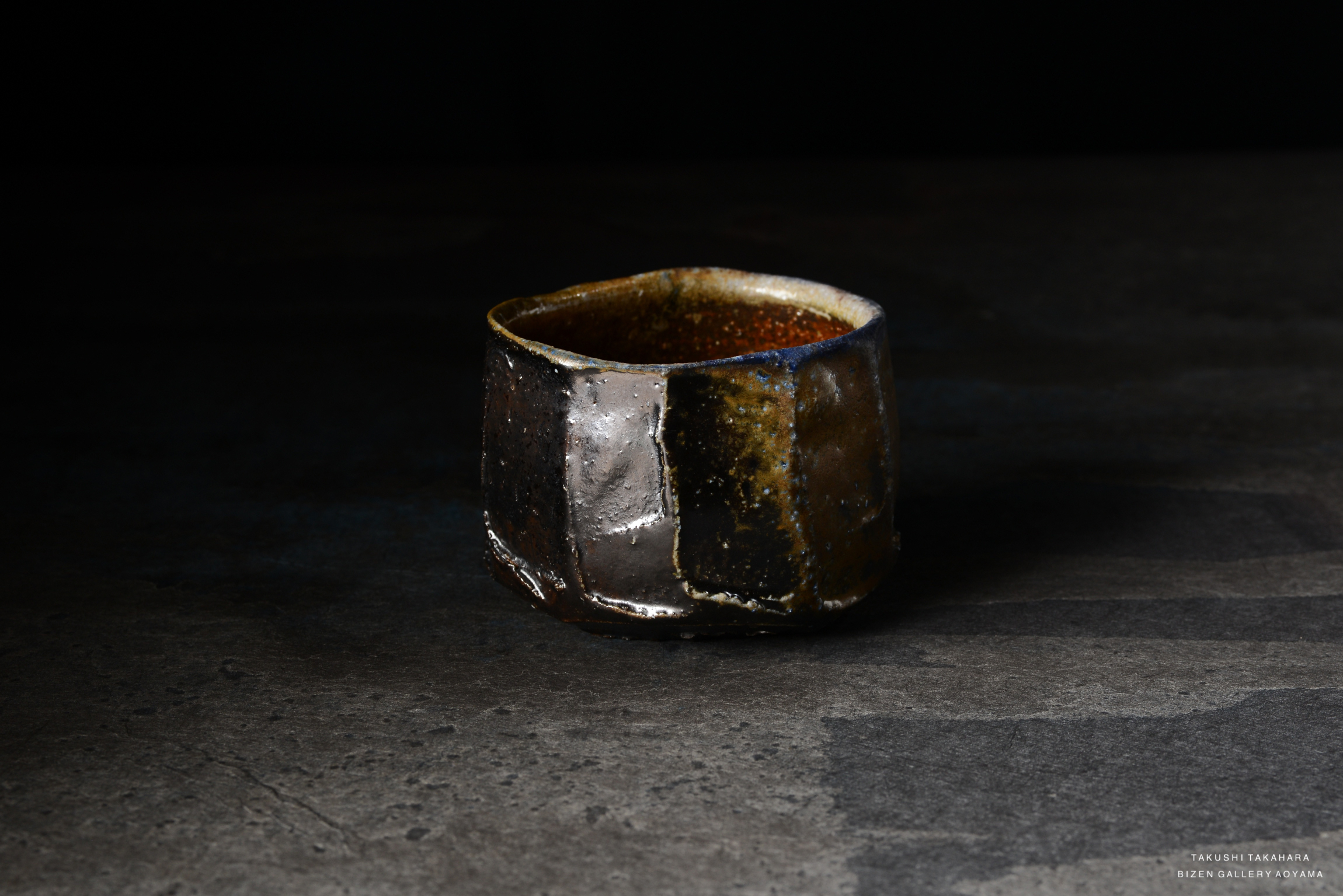 A handcrafted ceramic teacup with a unique, textured surface, featuring earthy tones of brown, gold, and blue, resting on a dark surface.