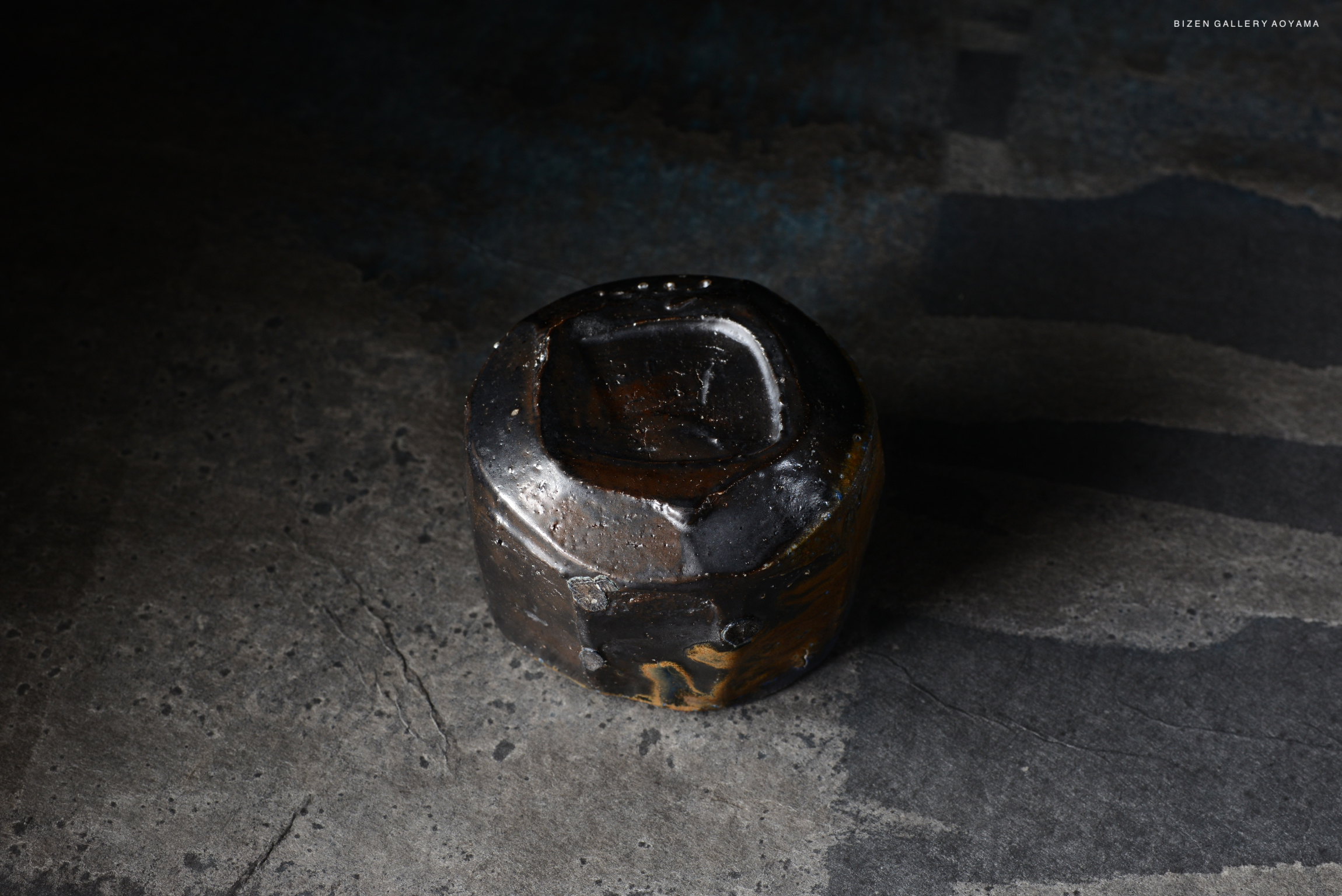 A unique, abstract ceramic piece with a dark, textured surface, displayed on a concrete background.