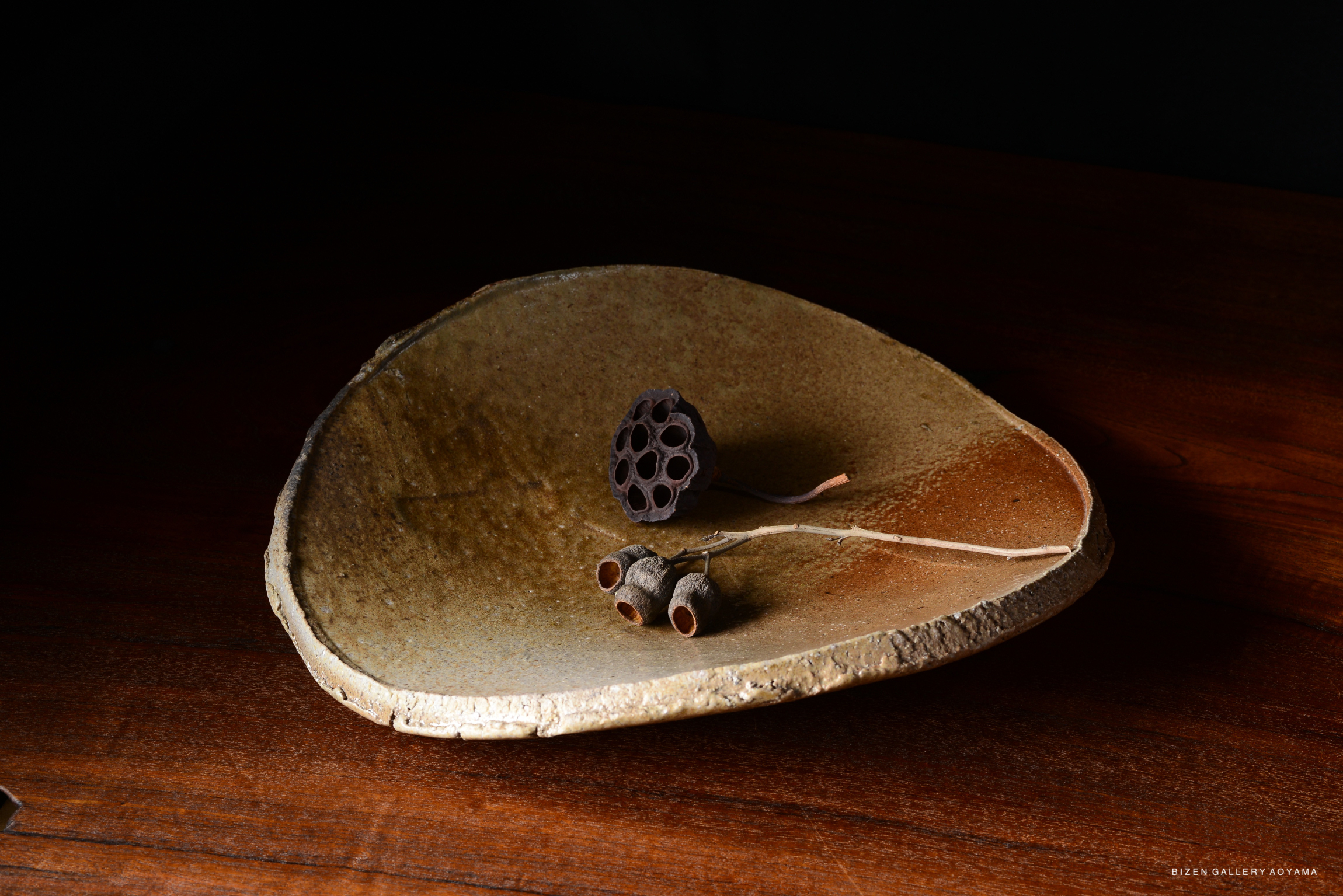 A rustic, oval-shaped ceramic plate with a textured surface, adorned with a dried lotus seed pod and two small, dried seed capsules on a wooden surface.