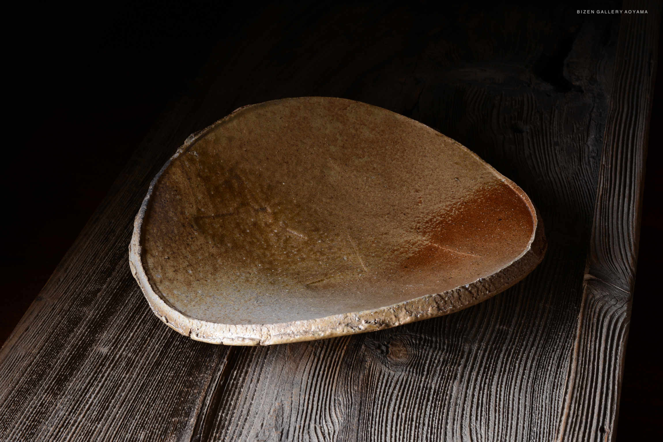 A close-up view of a large, earthy-toned ceramic plate sitting on a wooden surface, showcasing its organic shape and texture.
