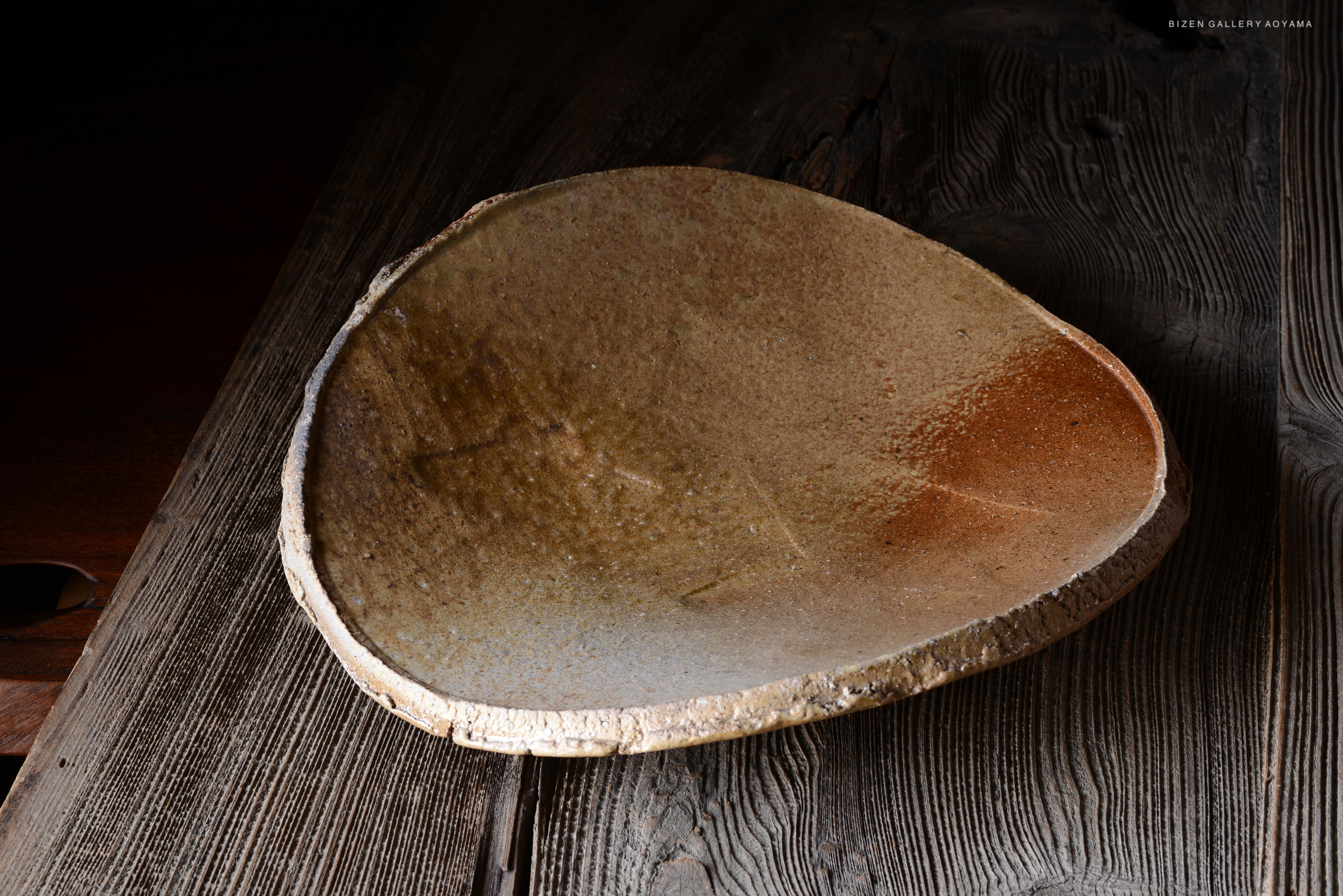 A close-up view of a rustic, oval-shaped ceramic bowl resting on a wooden surface, showcasing its textured, earthy glaze and natural edges.