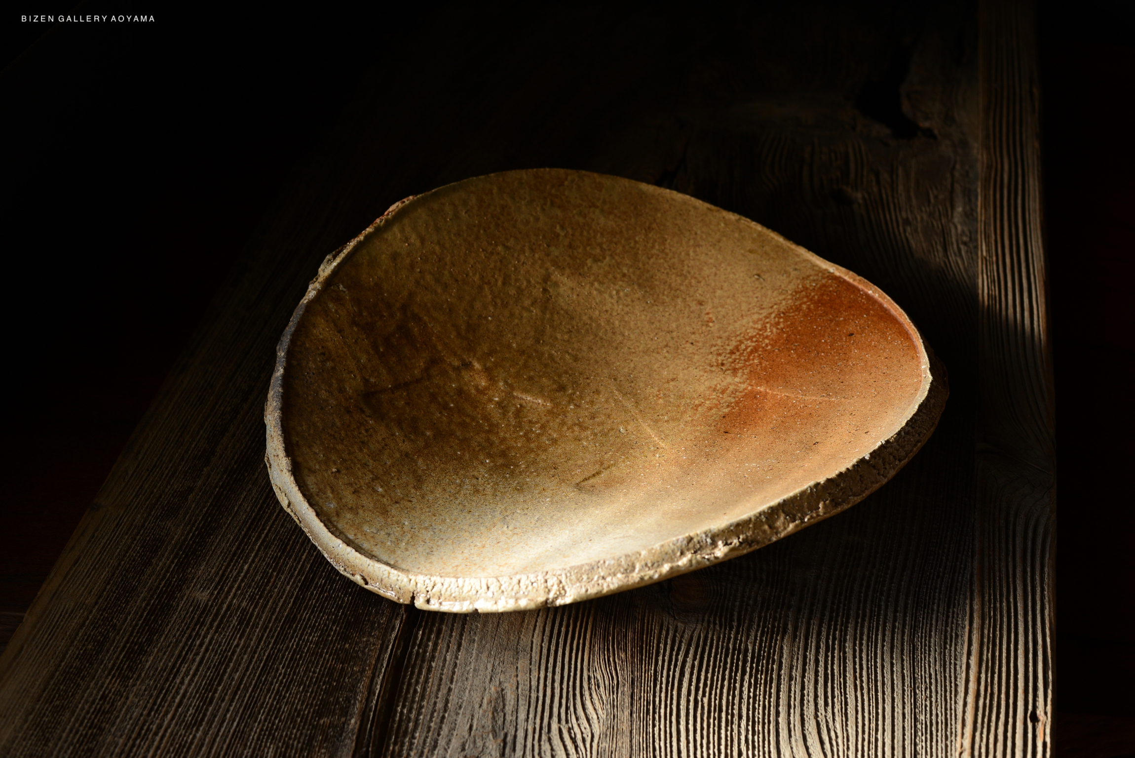 A close-up of a unique, handcrafted ceramic plate with a smooth, earthy texture, set against a dark background with wooden surfaces.