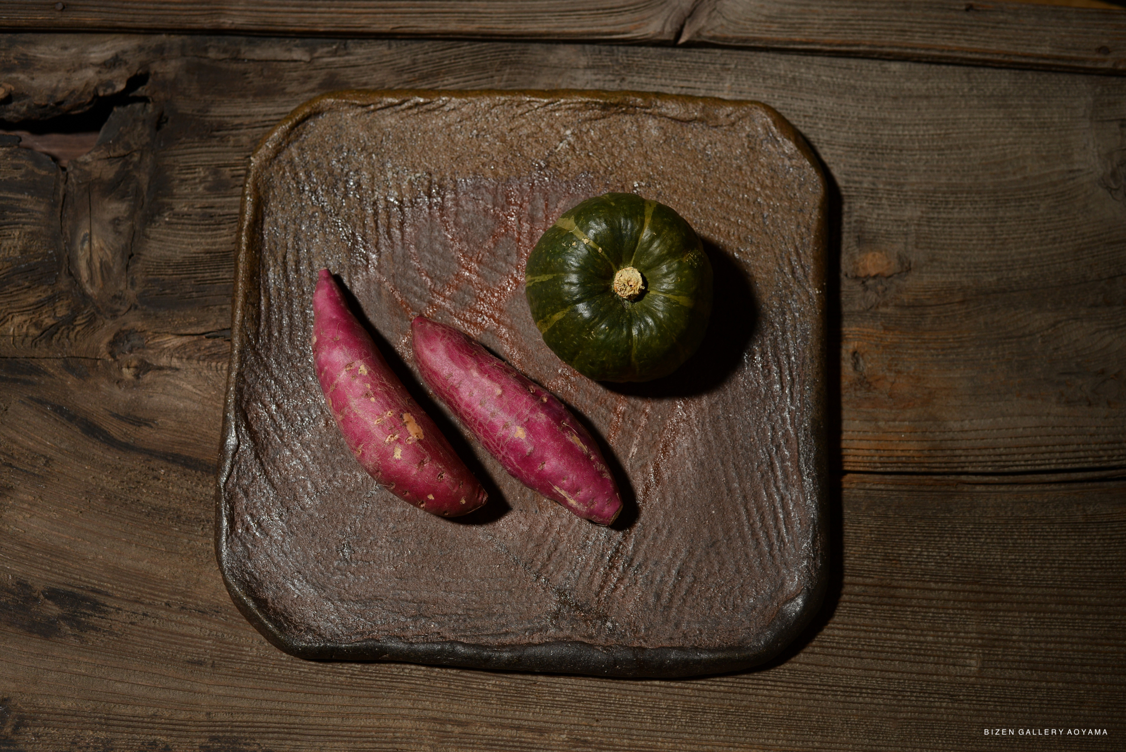 A rustic plate containing two purple sweet potatoes and a small green pumpkin, set against a wooden surface.