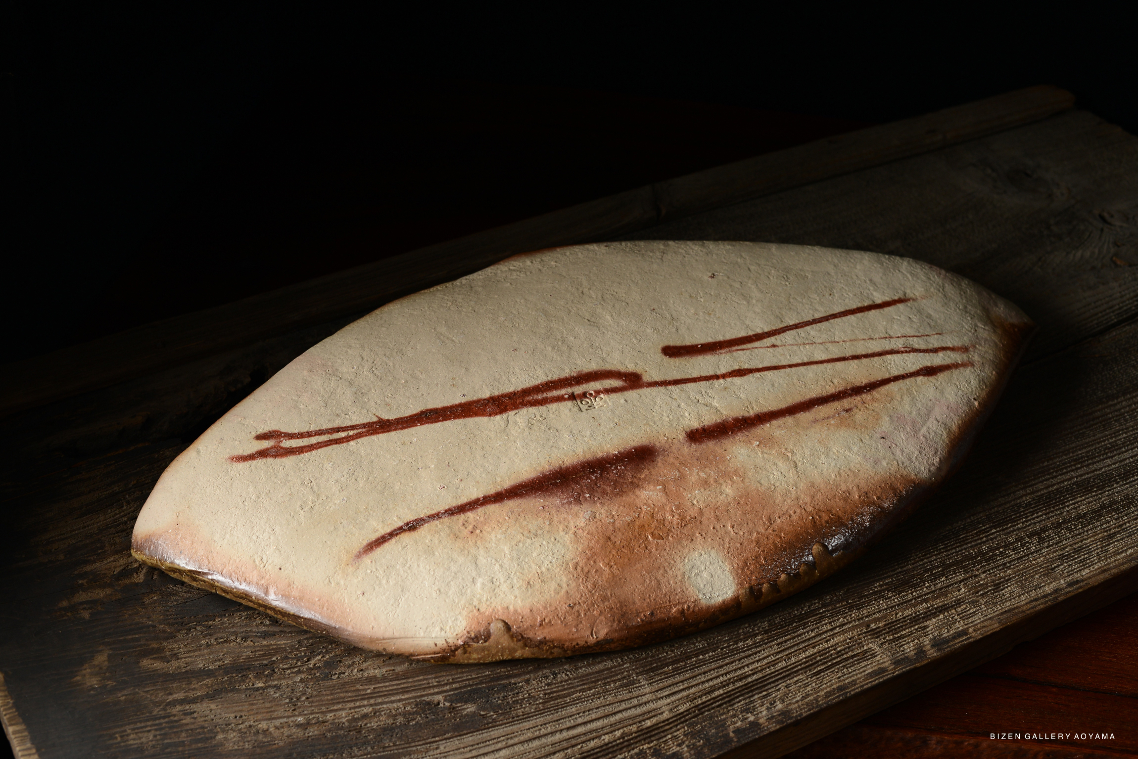 A close-up view of a flat, oval-shaped piece of pottery with a textured surface and red streaks, resting on a wooden surface.