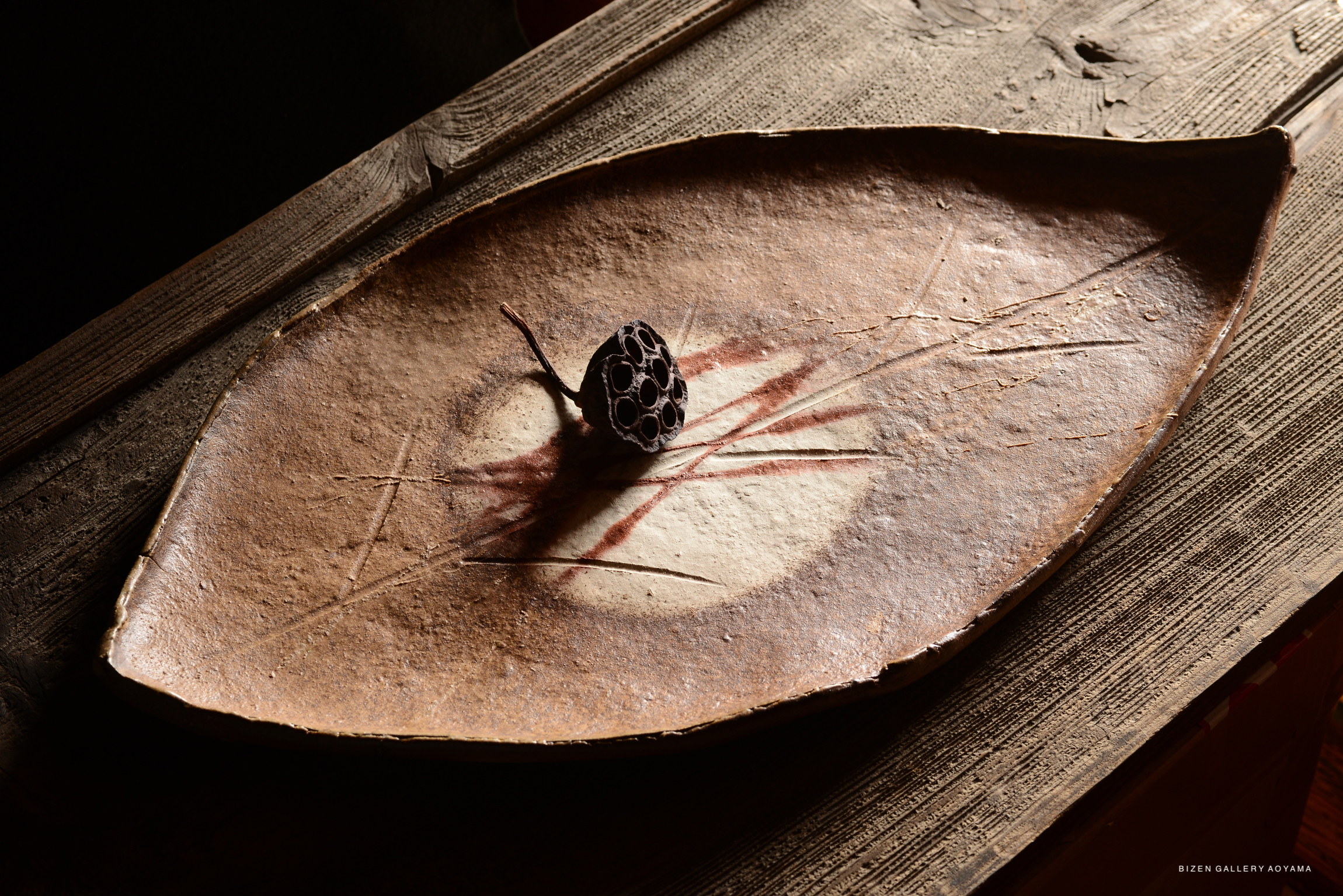 A ceramic platter in the shape of a leaf, featuring a decorative lotus seed pod placed in the center, set on a rustic wooden surface.