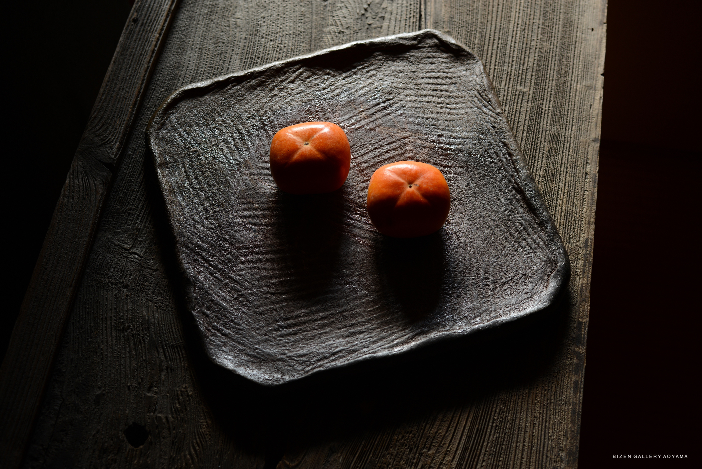 A dark, textured ceramic plate with two round, orange persimmons on top, set against a wooden surface.