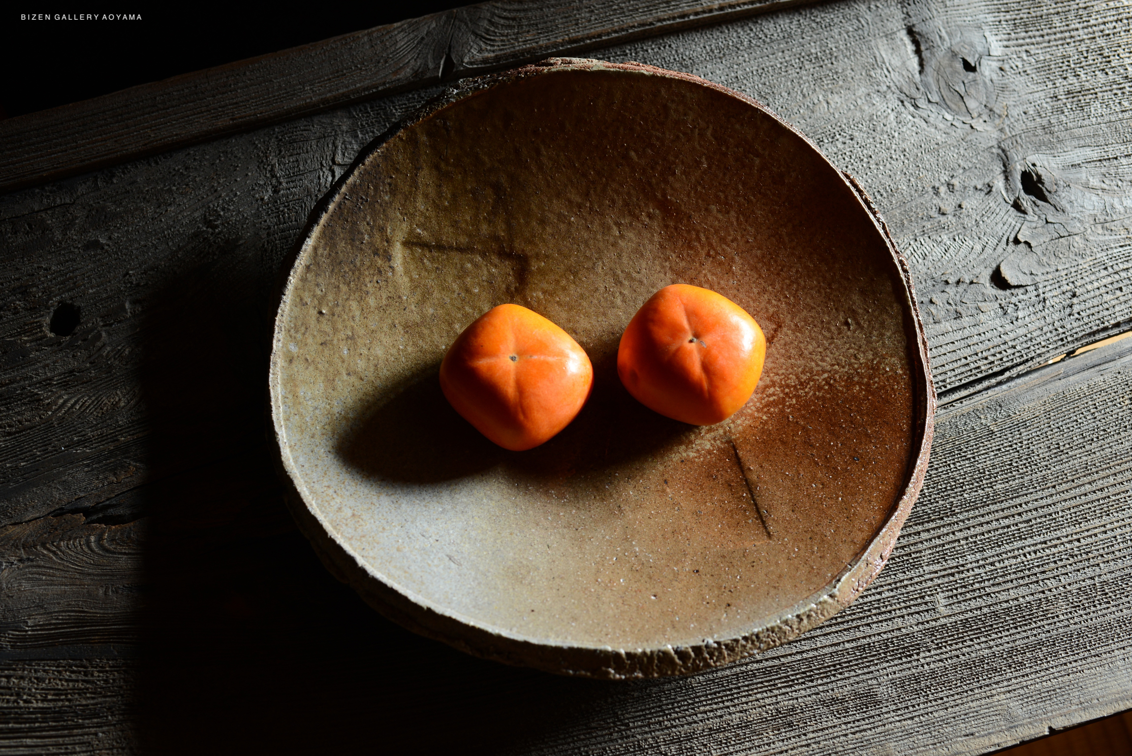 A rustic ceramic plate with two vibrant orange persimmons on a wooden surface.