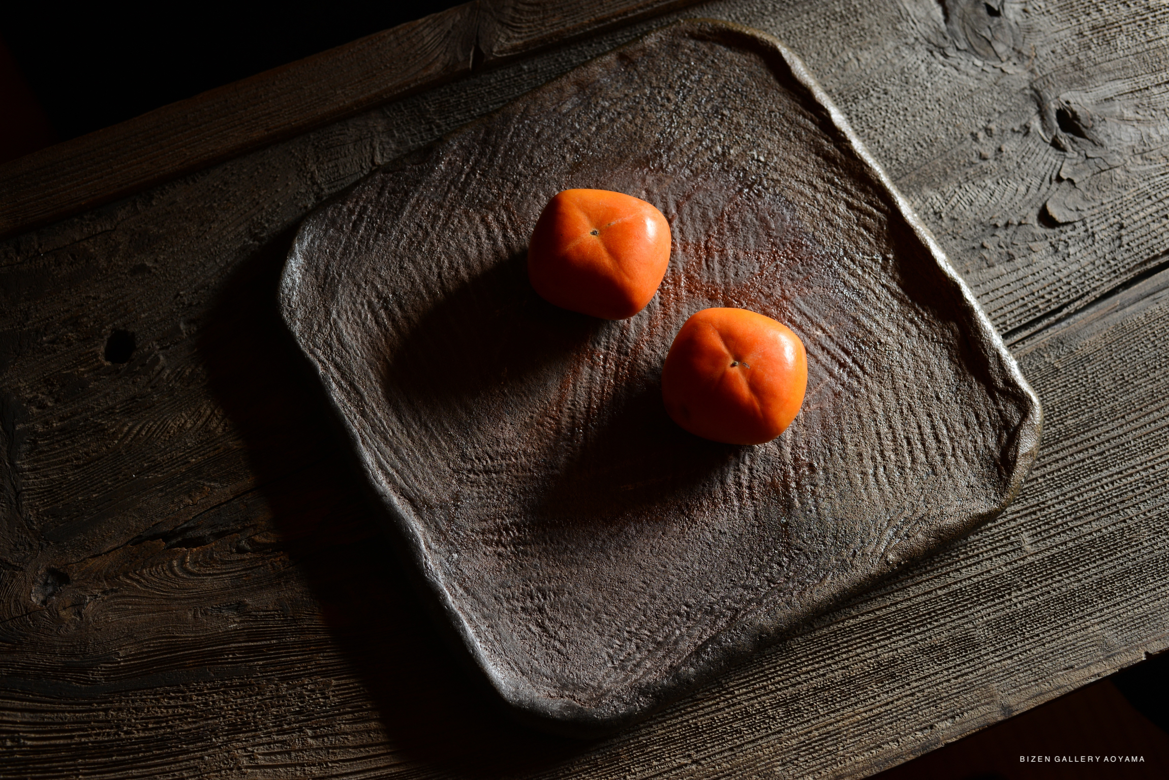 A close-up image of a rustic, dark brown slab plate with two bright orange persimmons placed on it. The background features a textured wooden surface, creating a warm and natural atmosphere.