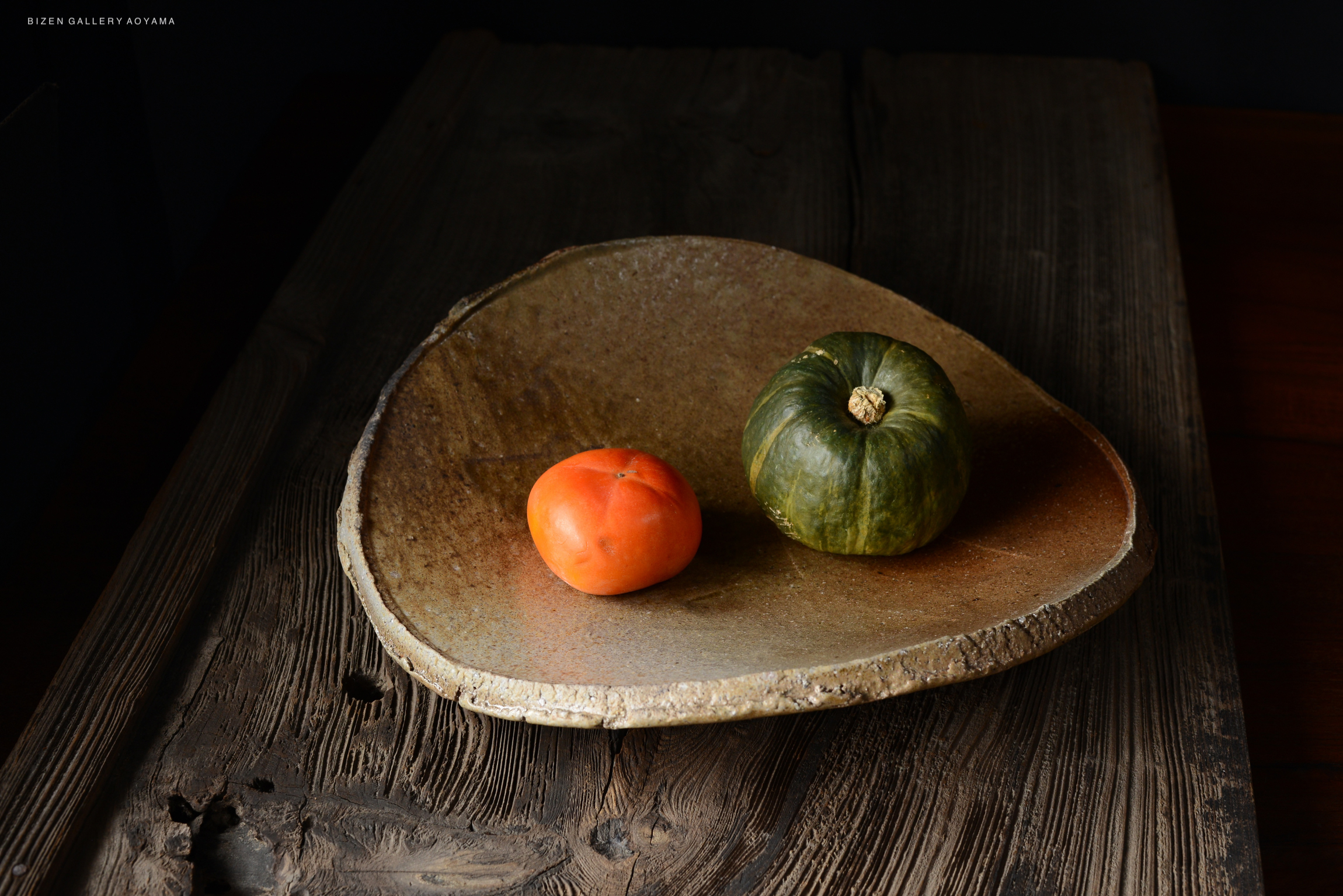 A rustic platter made of natural clay, displaying a vibrant orange persimmon and a green pumpkin, set against a dark wooden background.