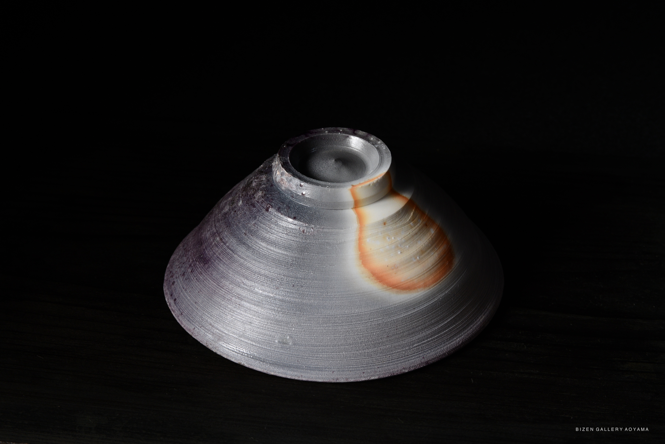 A Bizen pottery vessel with a conical shape, displaying a smooth texture and gradient color from purple to white, set against a dark background.