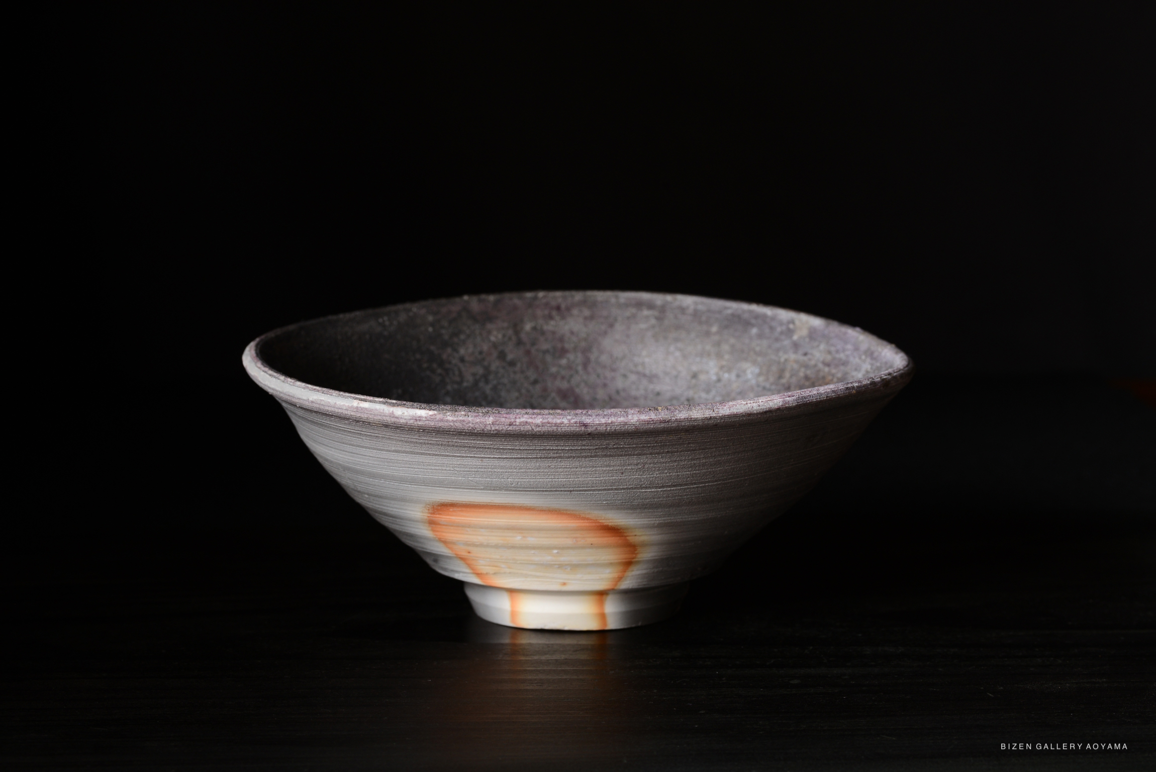 A handcrafted ceramic bowl with a textured gray surface and an orange accent at the base, set against a dark background.