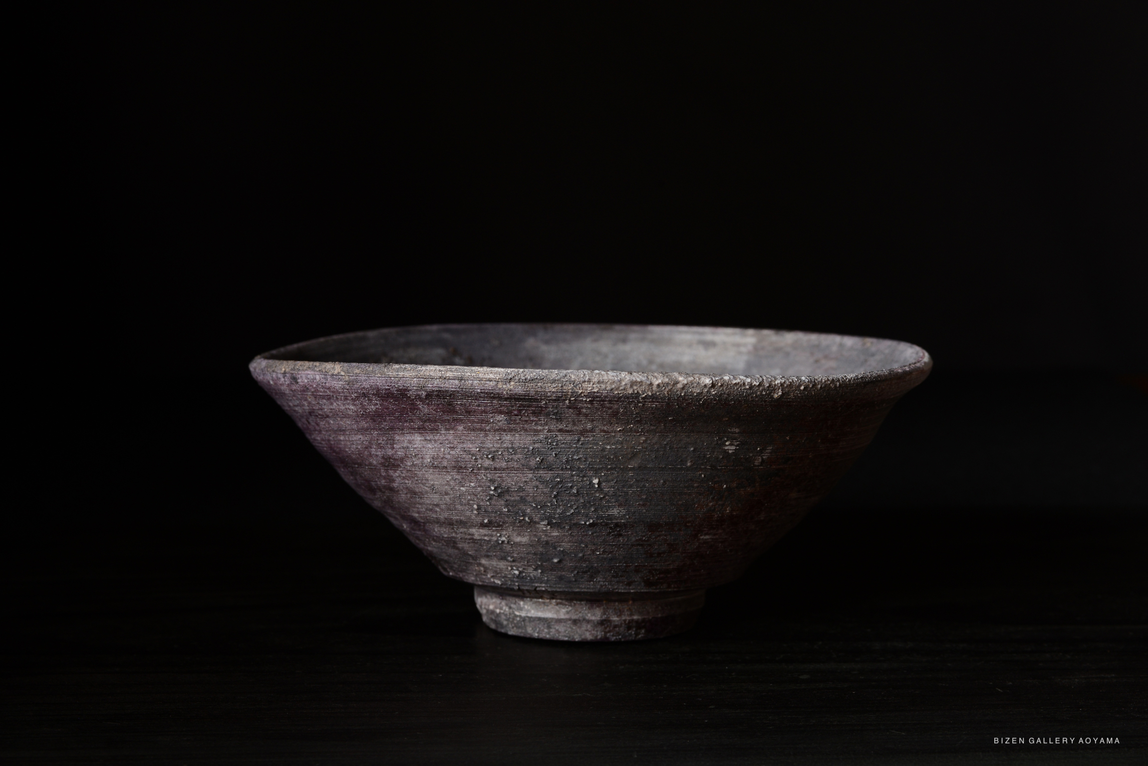 A handmade Bizen pottery bowl with a slightly flared rim and a textured surface, set against a dark background.