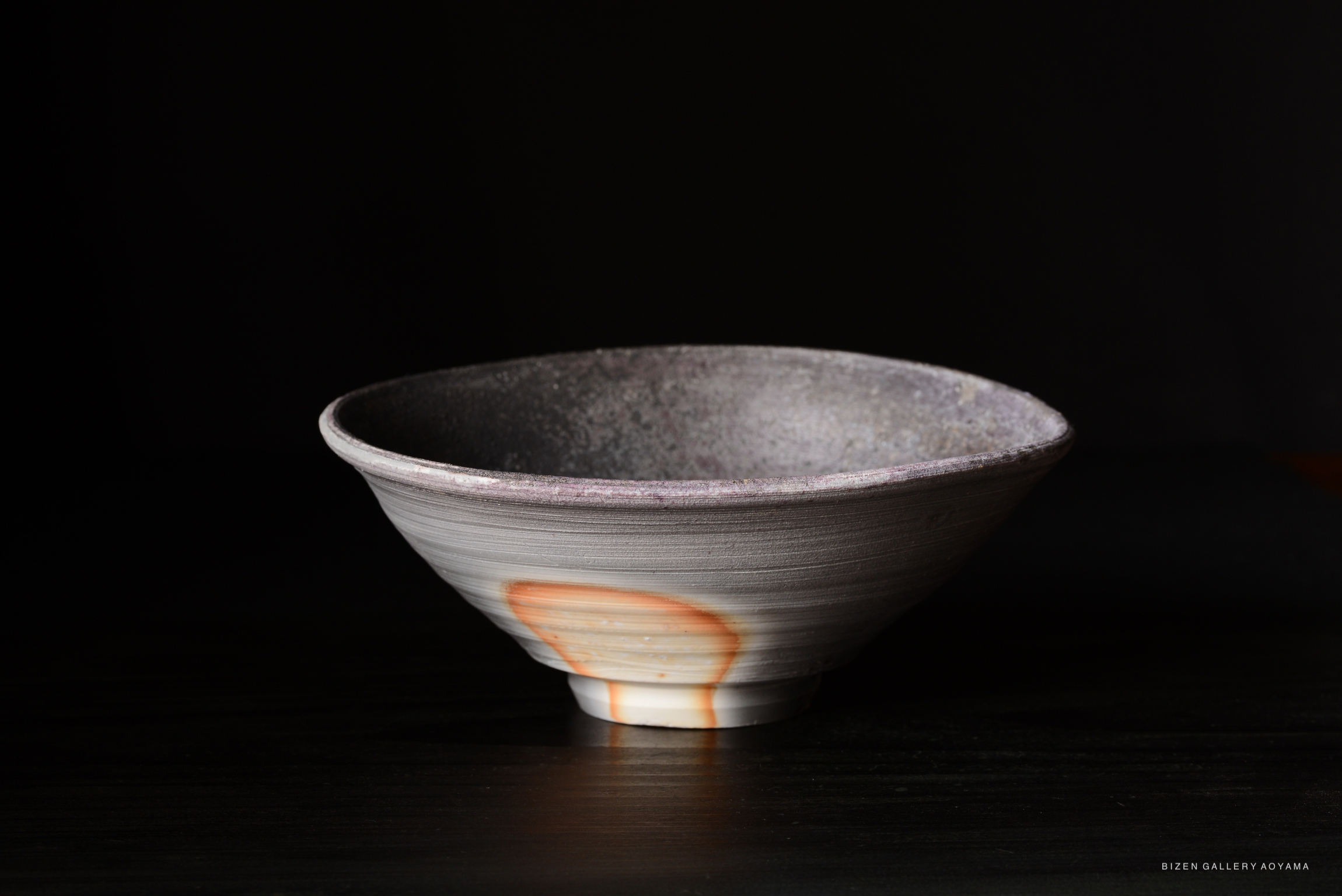 A ceramic bowl with a unique texture and a subtle gradient from gray to light color, featuring a distinctive mark or stain on the side, placed against a dark background.