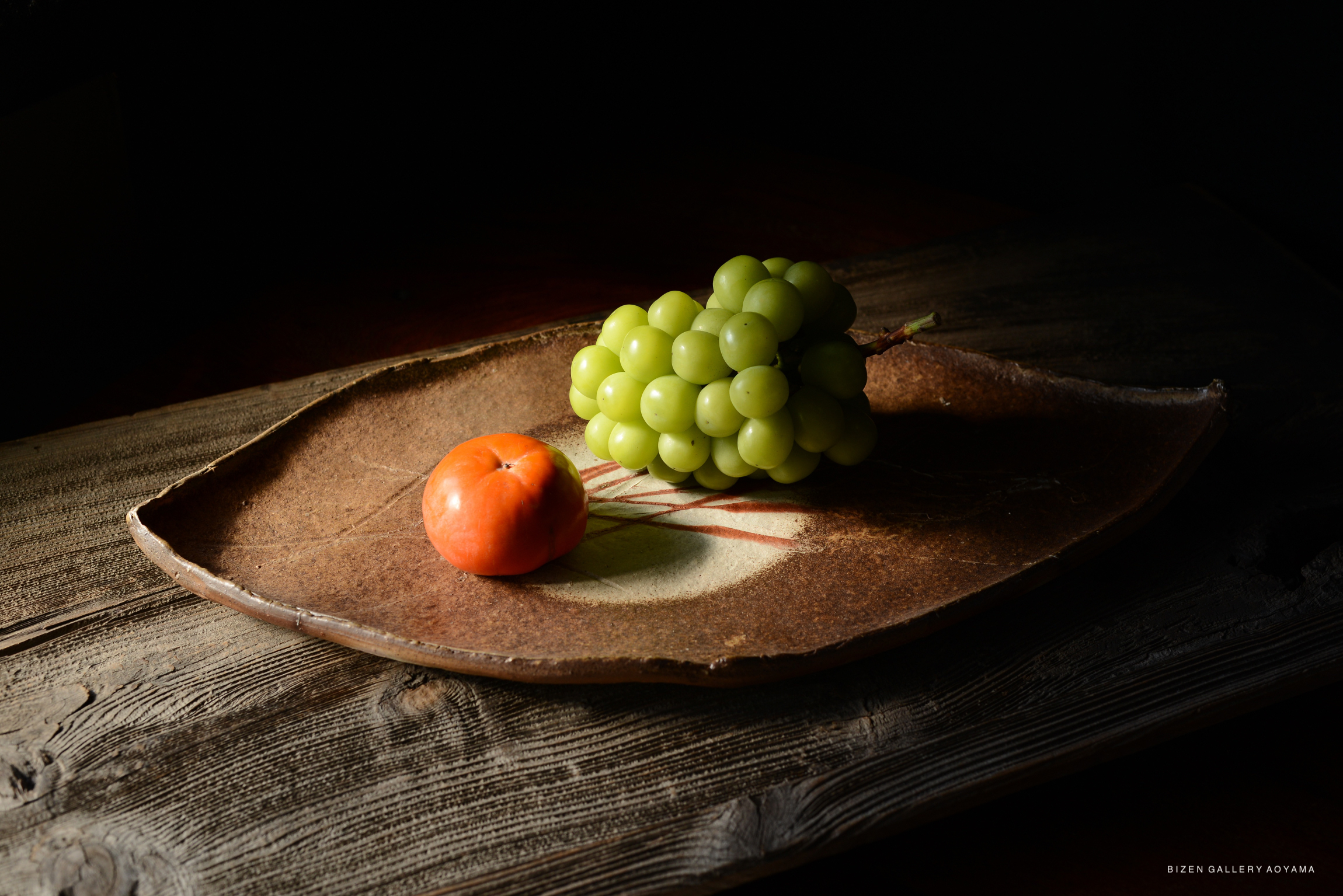 A leaf-shaped plate with a branch of green grapes and a persimmon arranged on it, set against a dark background.