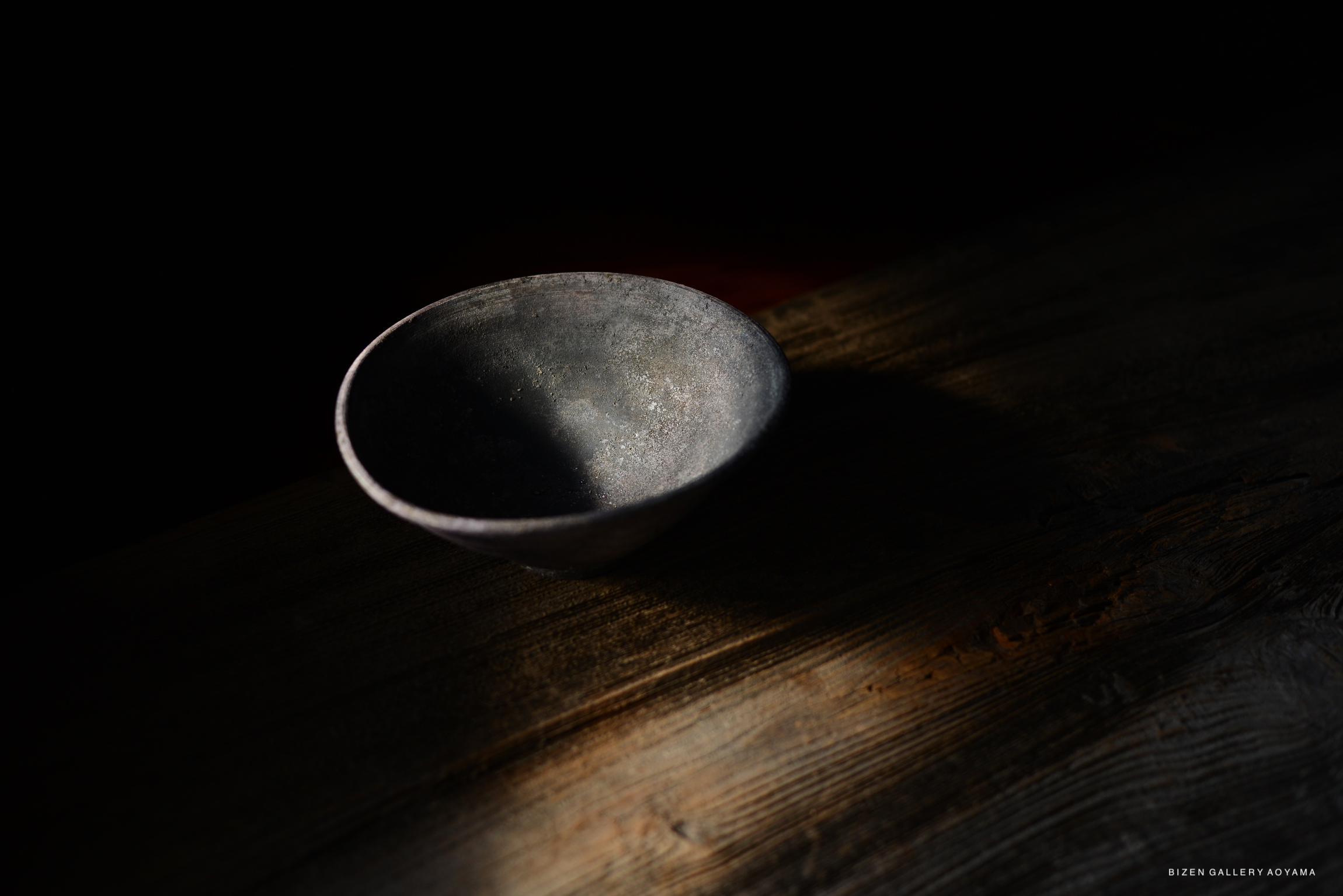 A rustic, dark ceramic bowl is placed on a wooden surface, illuminated by a patch of light, creating a contrast with the surrounding darkness.