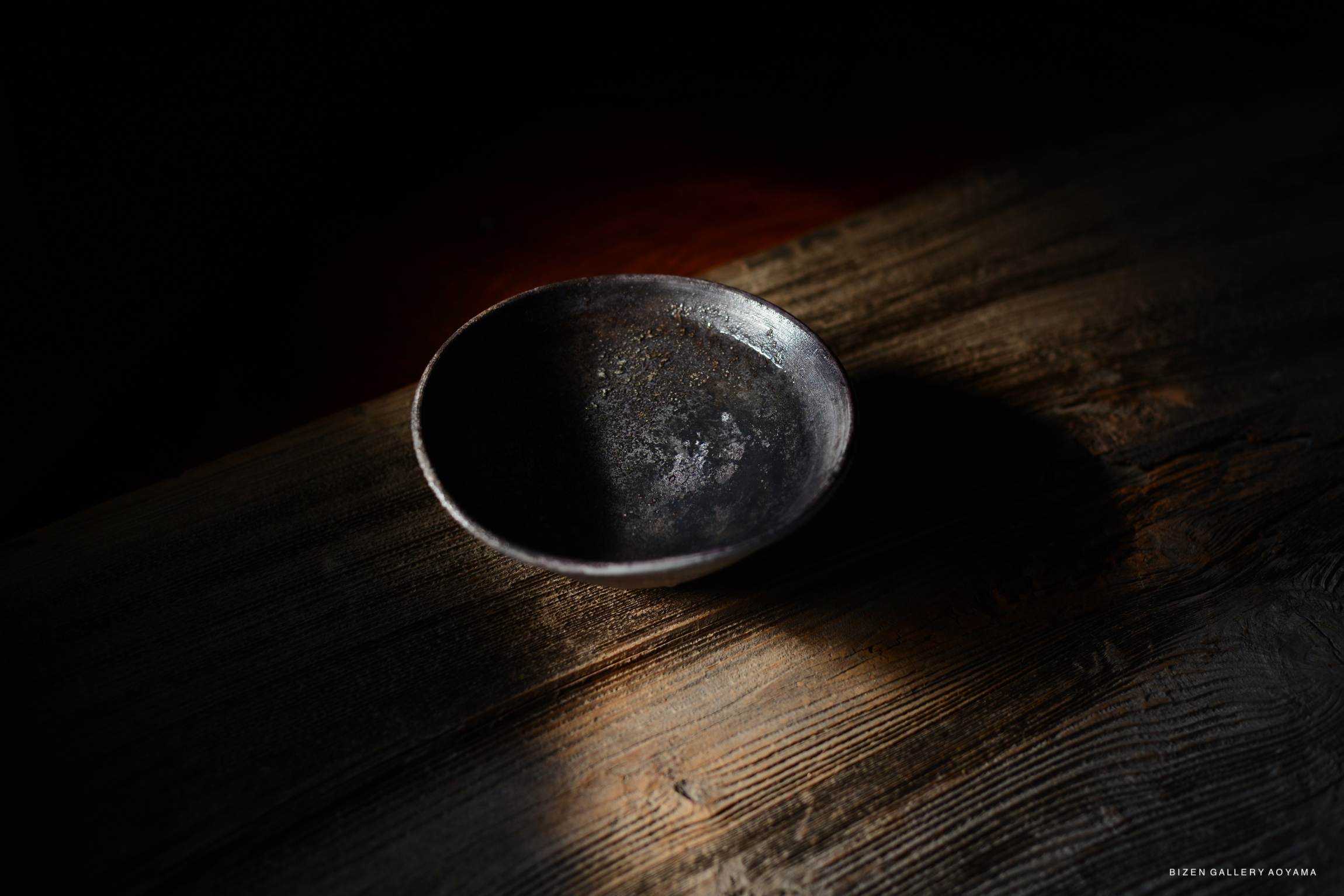 A dark ceramic bowl placed on a wooden surface, illuminated by soft light, creating elegant shadows.