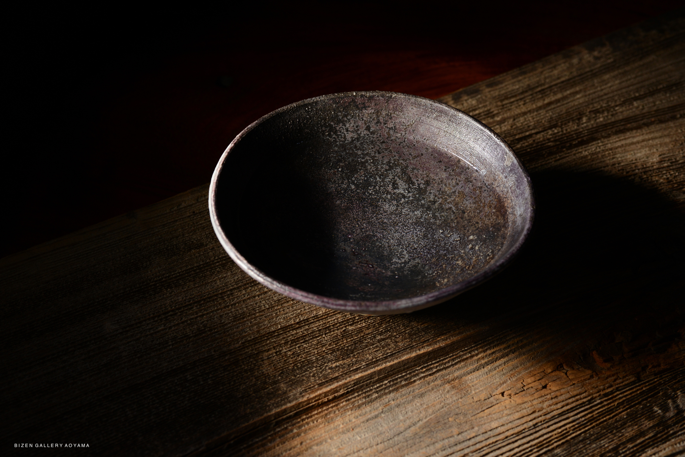 A close-up view of a dark, textured ceramic bowl placed on a rustic wooden surface, with soft shadows creating a dramatic effect.