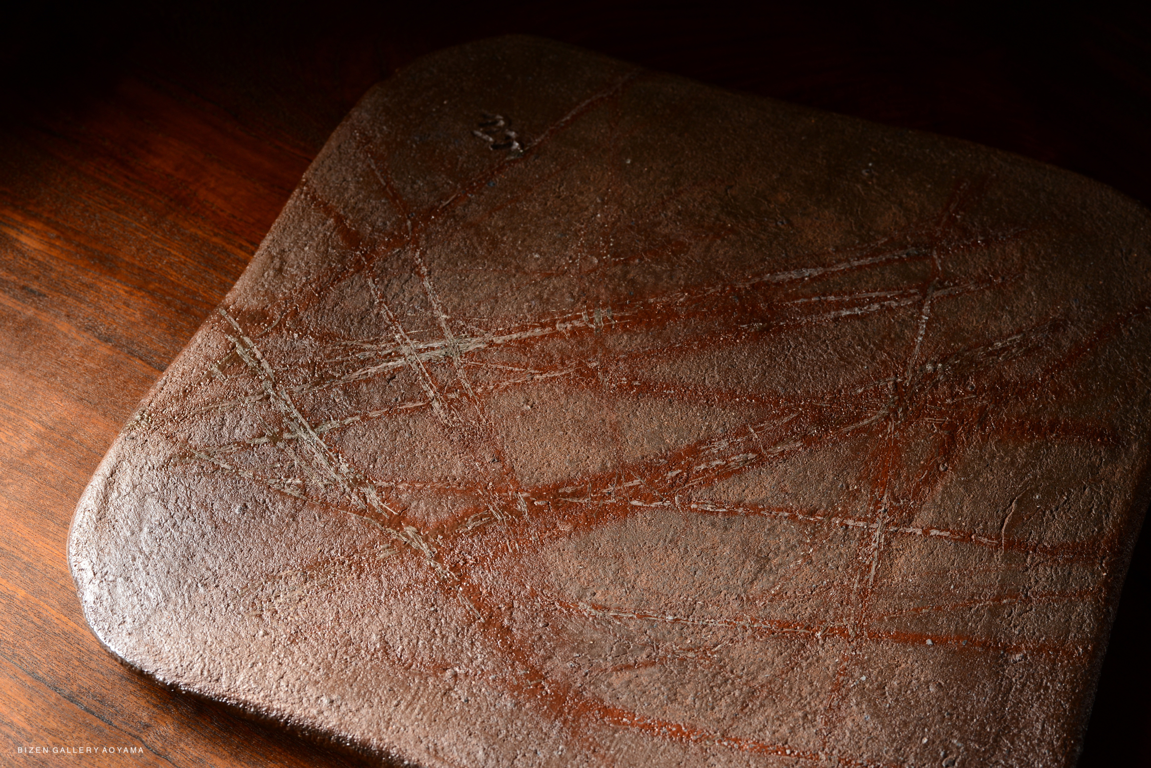 Close-up of a textured, dark brown ceramic plate with subtle markings and scratches on its surface, set against a wooden background.